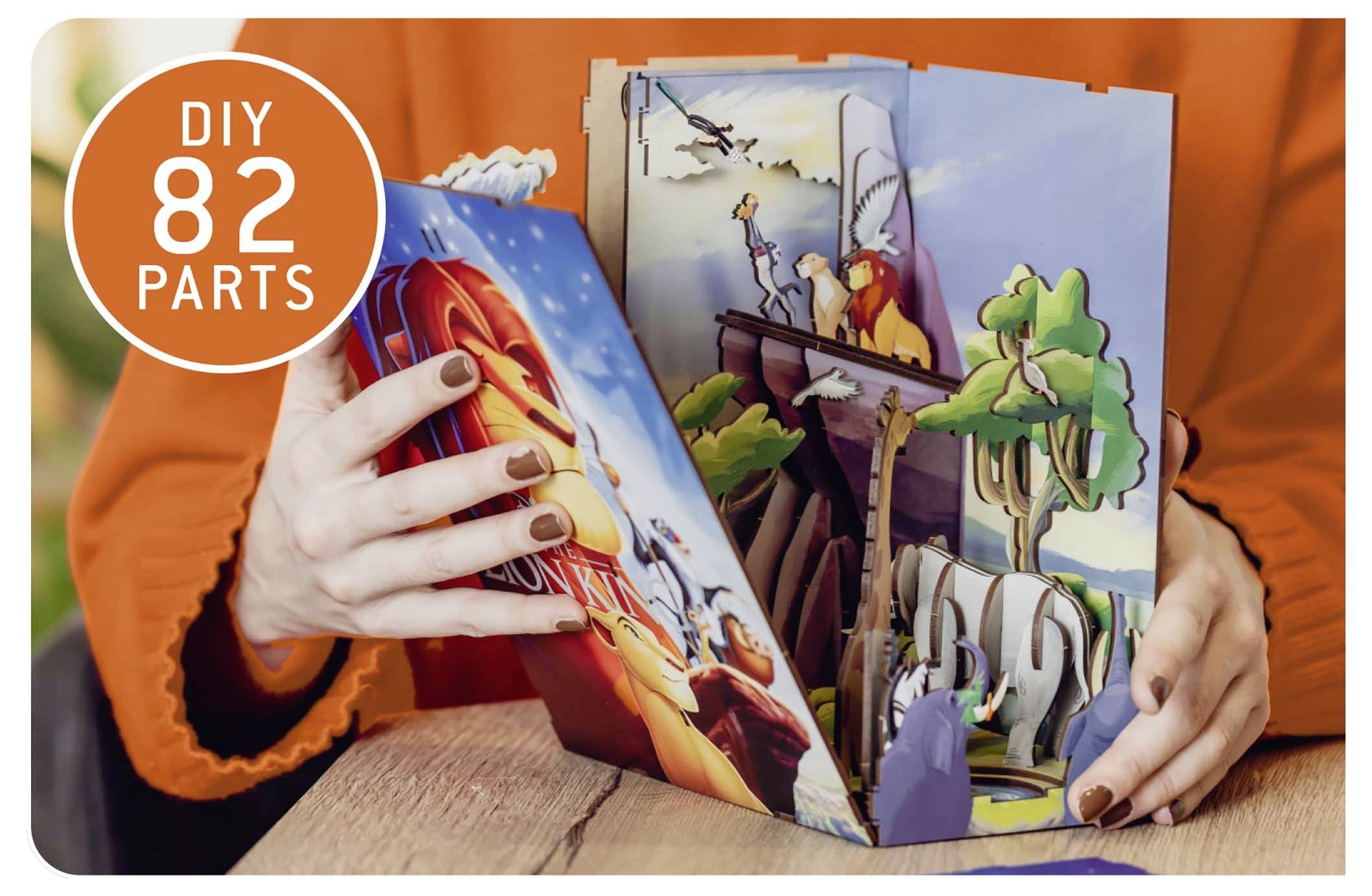 En person sätter ihop en färggrann DIY-popup-bok med texten 'DIY 82 delar'. Scenen visar djur och landskap i ett livligt diorama.