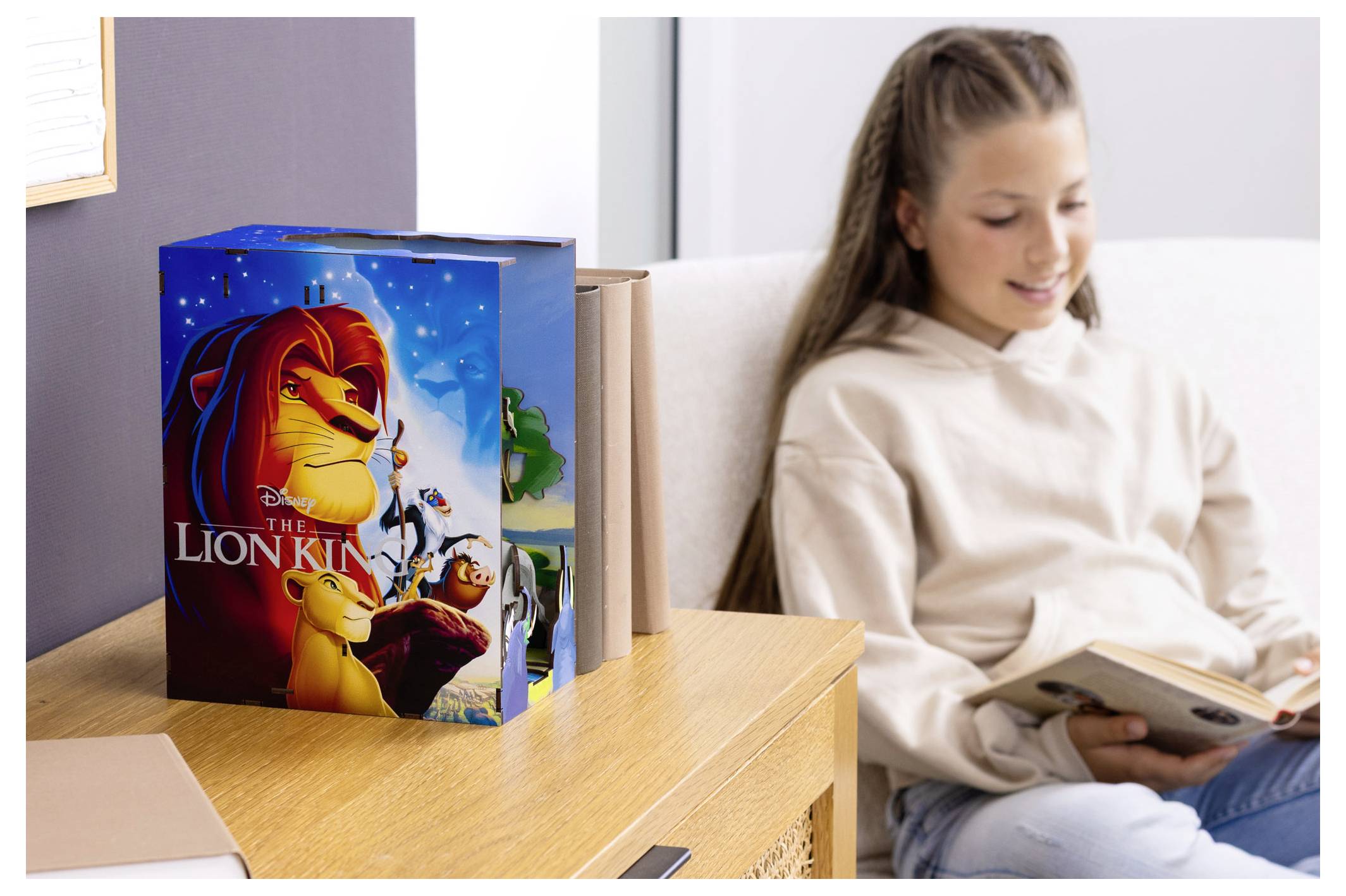 En ung flicka läser en bok bredvid en spelbox med 'Lejonkungen' konstverk som visar karaktärer. Rummet har en ljus, mysig atmosfär.