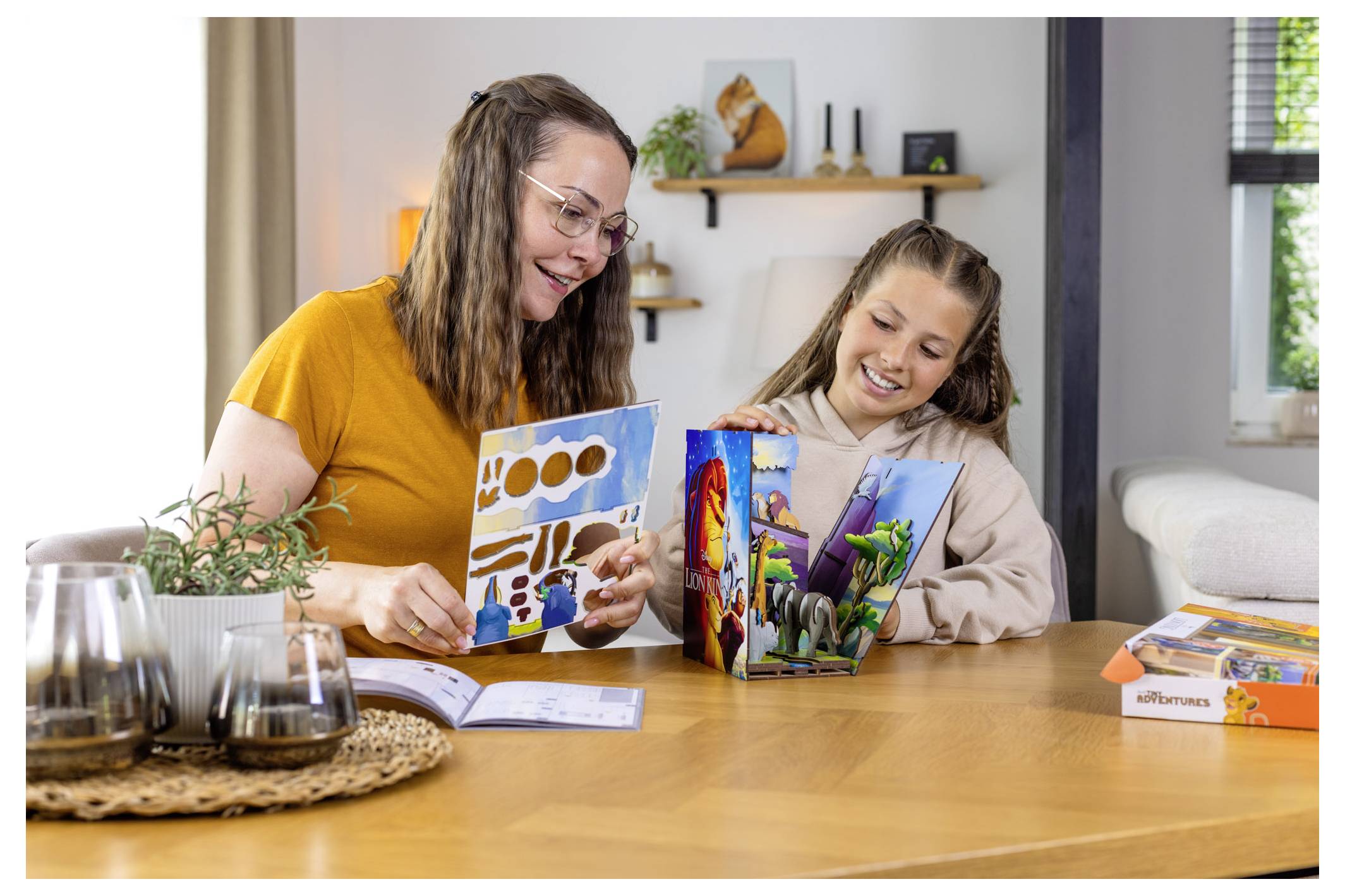 En kvinna och en ung flicka sitter vid ett bord och läser tillsammans i böcker med levande illustrationer, och de ler. Ett mysigt vardagsrum syns i bakgrunden.