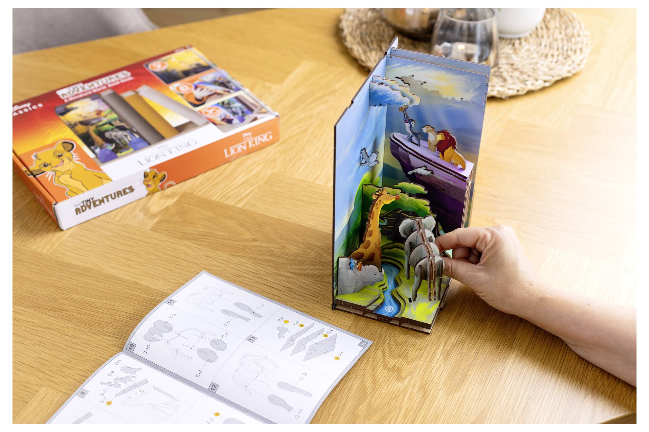 En person monterar ett 3D-pappersdiorama föreställande en scen med djur och en båt, bredvid en modellsats med tema från Lejonkungen och en instruktionsbok.