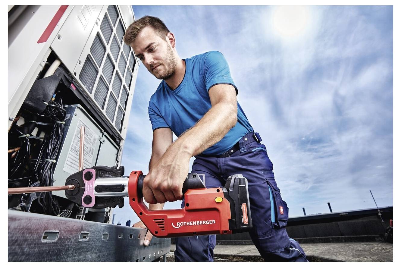 En tekniker i blå arbetsklädsel använder ett rött hydrauliskt verktyg för att arbeta på en stor maskin utomhus under en molnig himmel, med intensivt fokus.