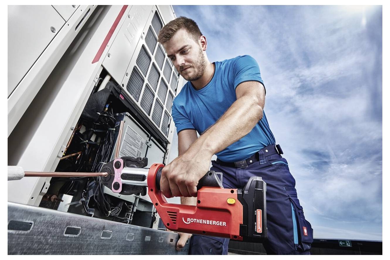 En tekniker använder ett rött verktyg märkt 'ROTHENBERGER' för att arbeta med elektrisk utrustning utomhus, iklädd en blå uniform under en molnig himmel.