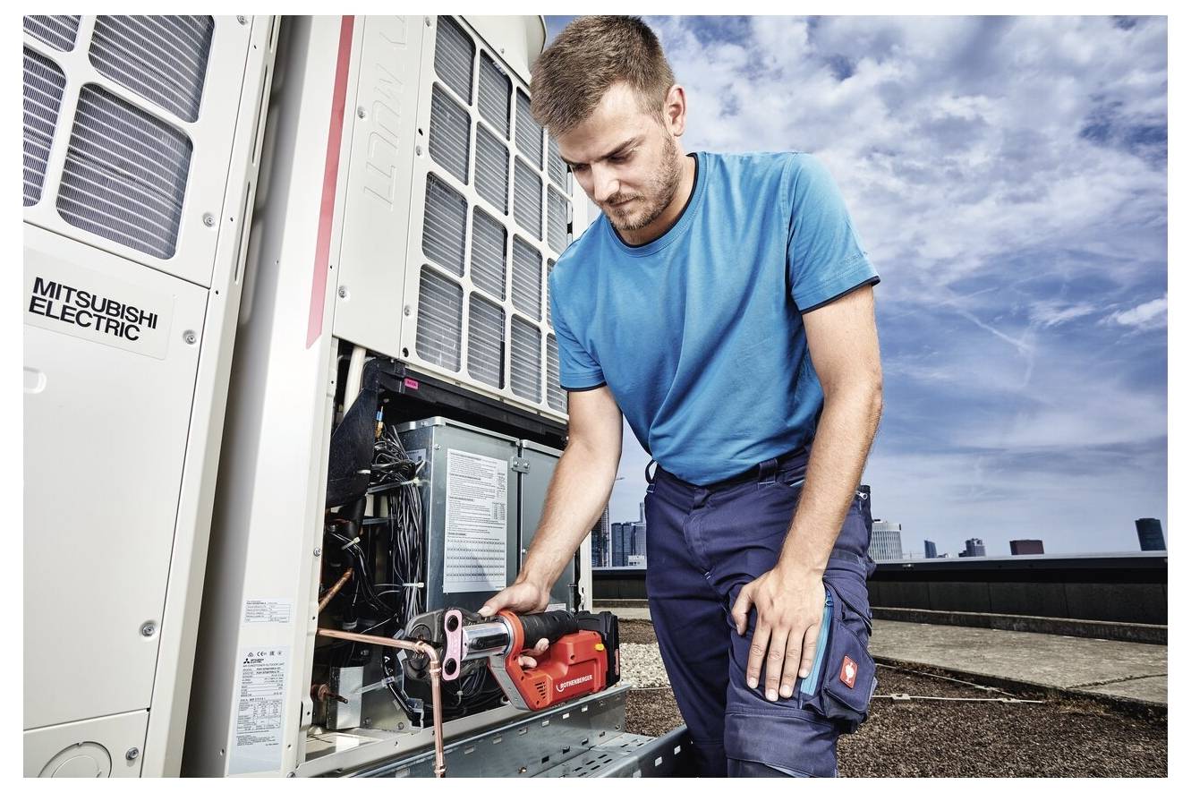 En tekniker i en blå skjorta och arbetsbyxor reparerar en industriell luftkonditioneringsenhet med ett verktyg på ett tak, med en stadssiluett i bakgrunden.