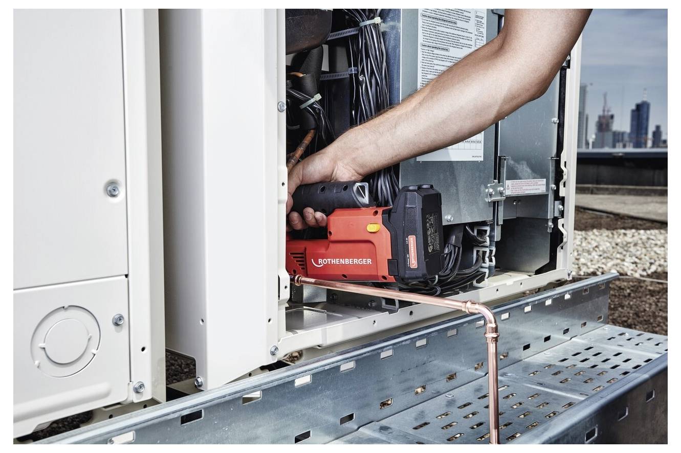 En person som använder ett rött Rothenberger-verktyg för att arbeta på en elinstallation på ett tak, med stadsbyggnader synliga i bakgrunden.