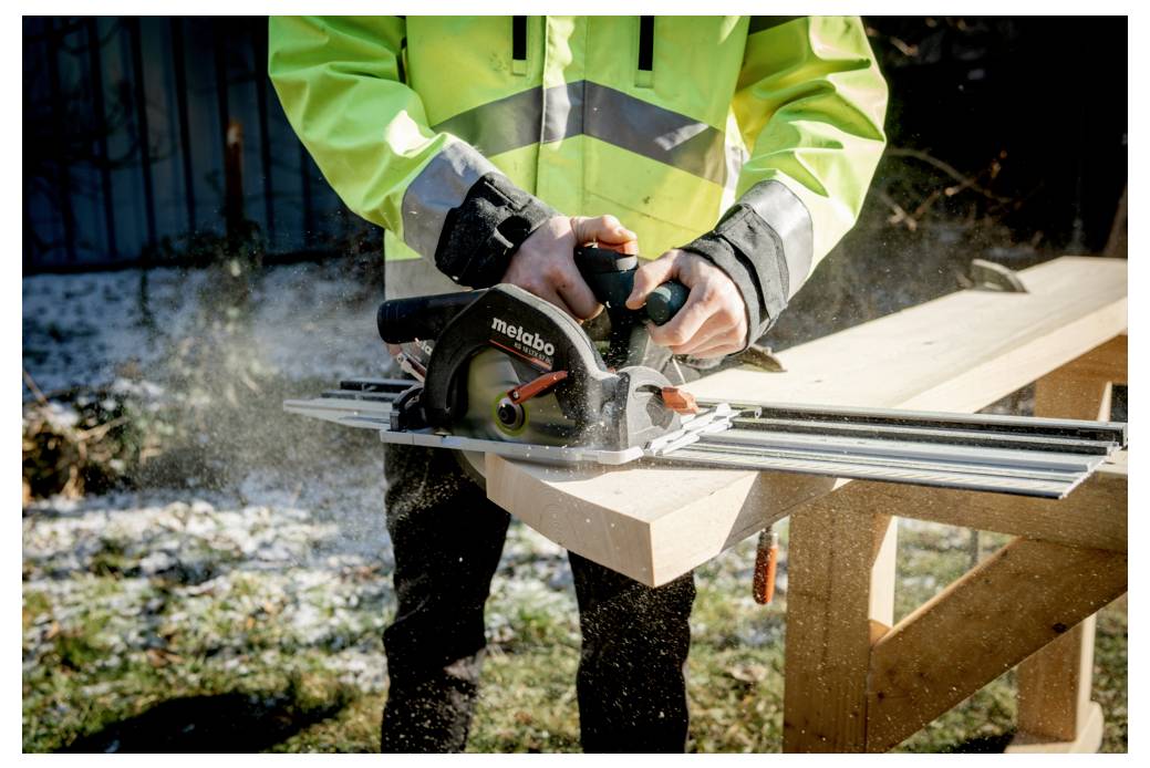 'En person som använder en Metabo cirkelsåg för att såga ett träplank utomhus, iklädd en ljusgrön reflexjacka och handskar.'