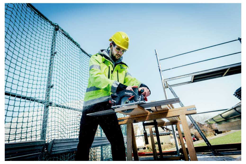 En byggnadsarbetare med skyddshjälm och varselväst använder en cirkelsåg för att såga träbalkar på en utomhusarbetsplats.