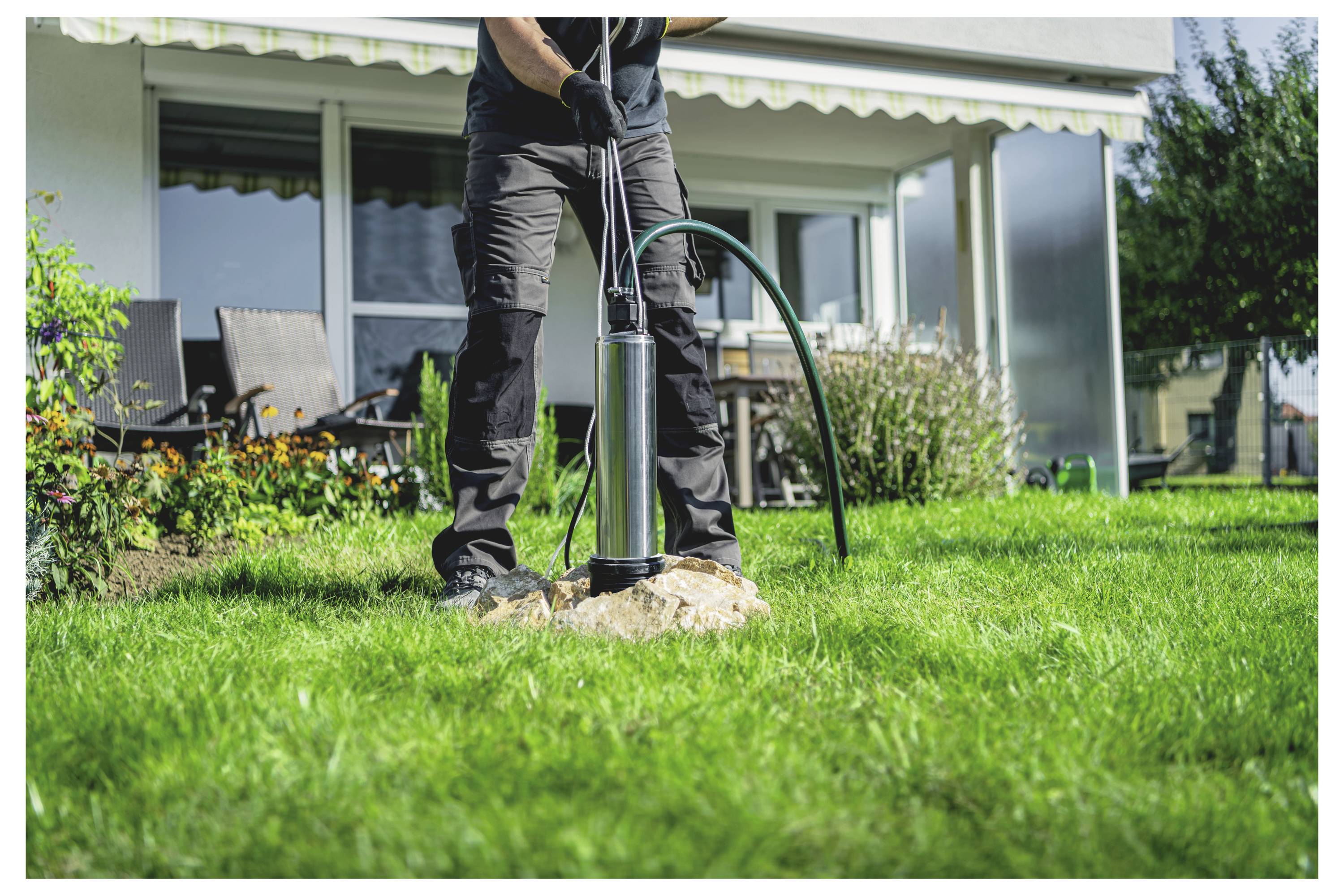 En person i en trädgård som använder en metallpump för att vattna växter, med ett hus och en uteplats i bakgrunden.
