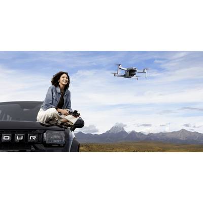 En kvinna sitter i en svart 4x4 SUV och håller i en drönarkontroll. En drönare flyger i närheten över ett vackert landskap med berg i bakgrunden.