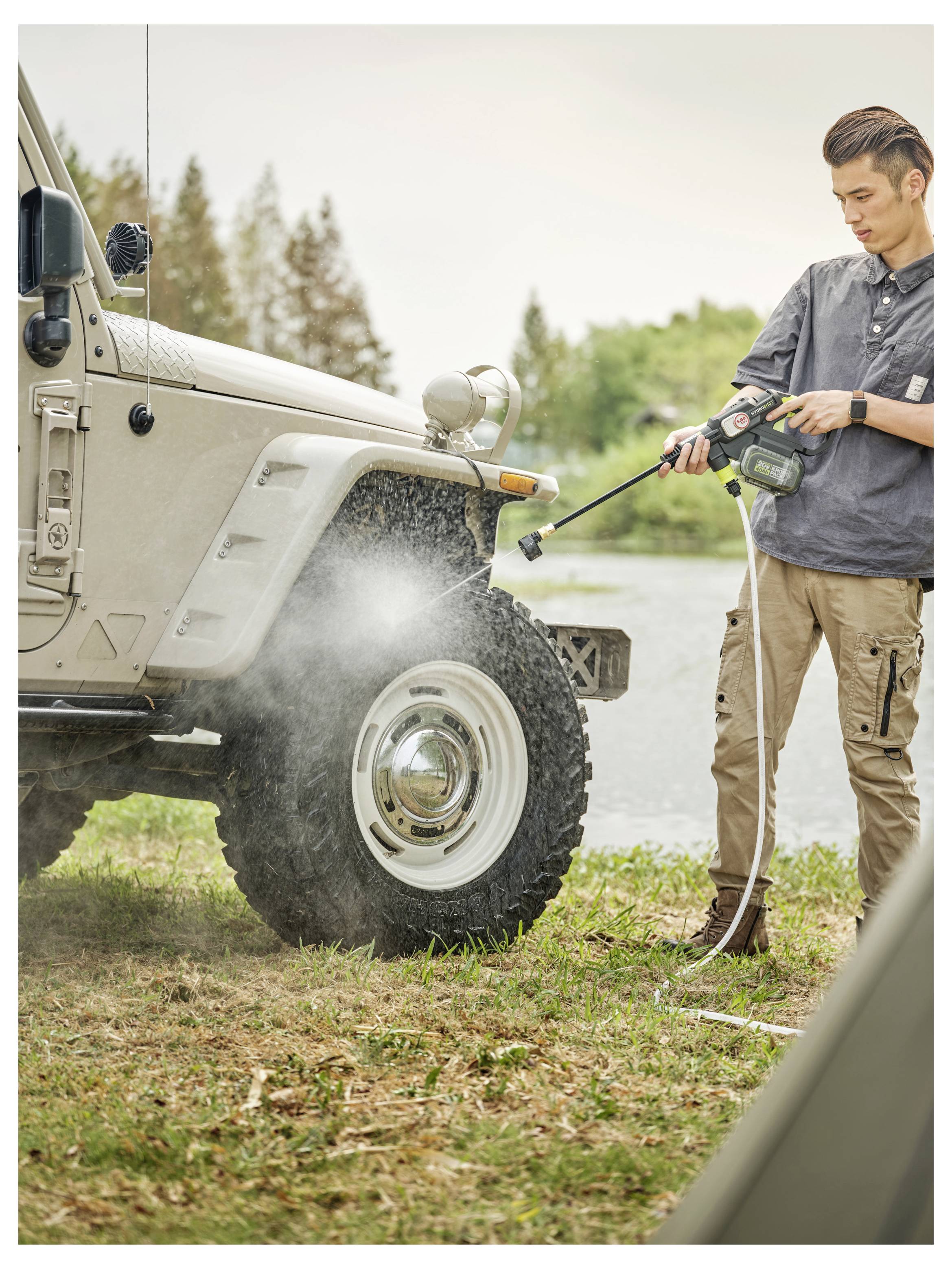 En man högtryckstvätta framhjulet och sidan på en beige Jeep Wrangler utomhus nära vatten och rensar lera från däcket.