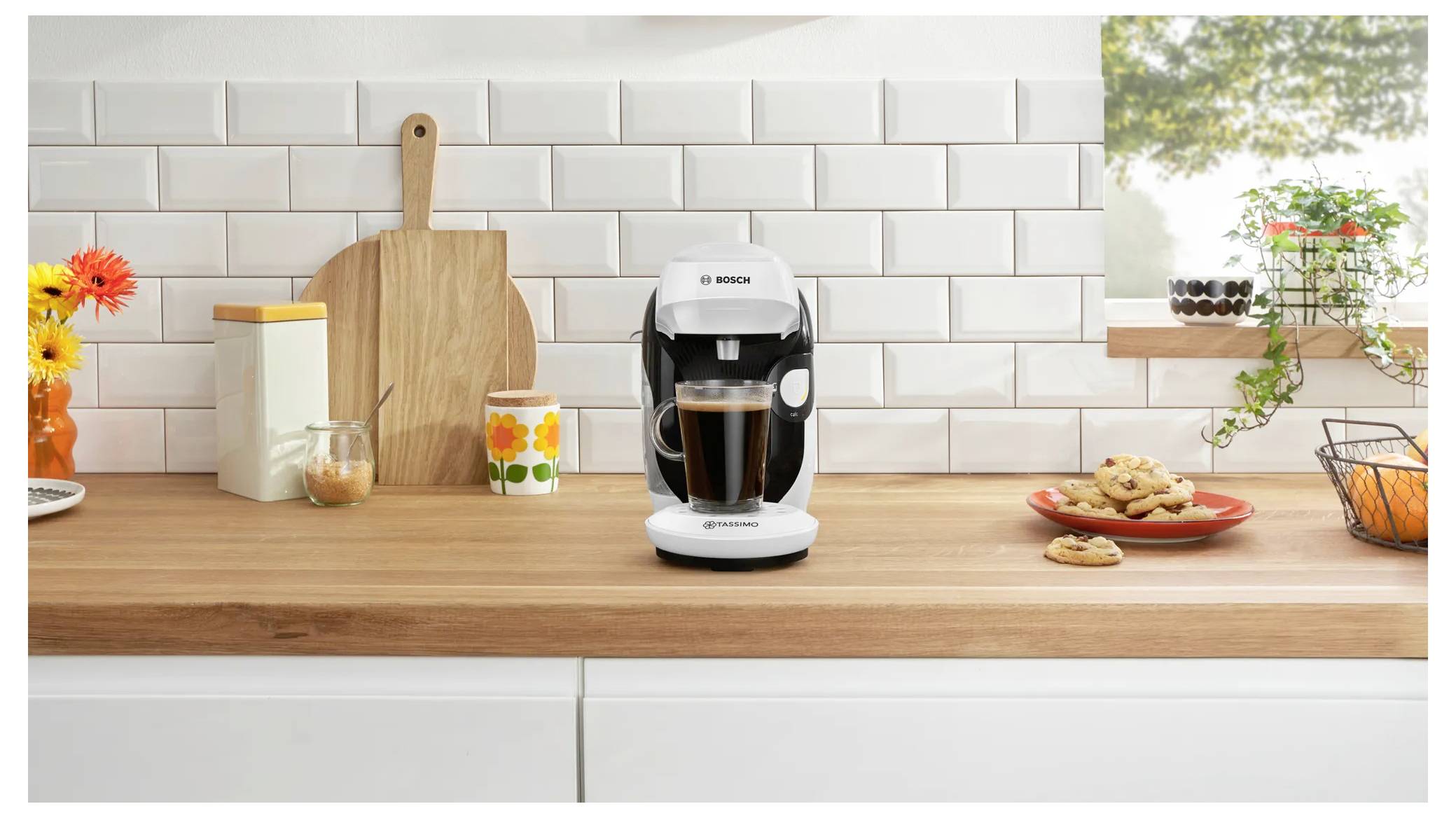 En modern kaffebryggare som brygger en kopp på en träköksbänk; omgiven av kakor, blommor och burkar mot en vit kaklad stänkskiva.