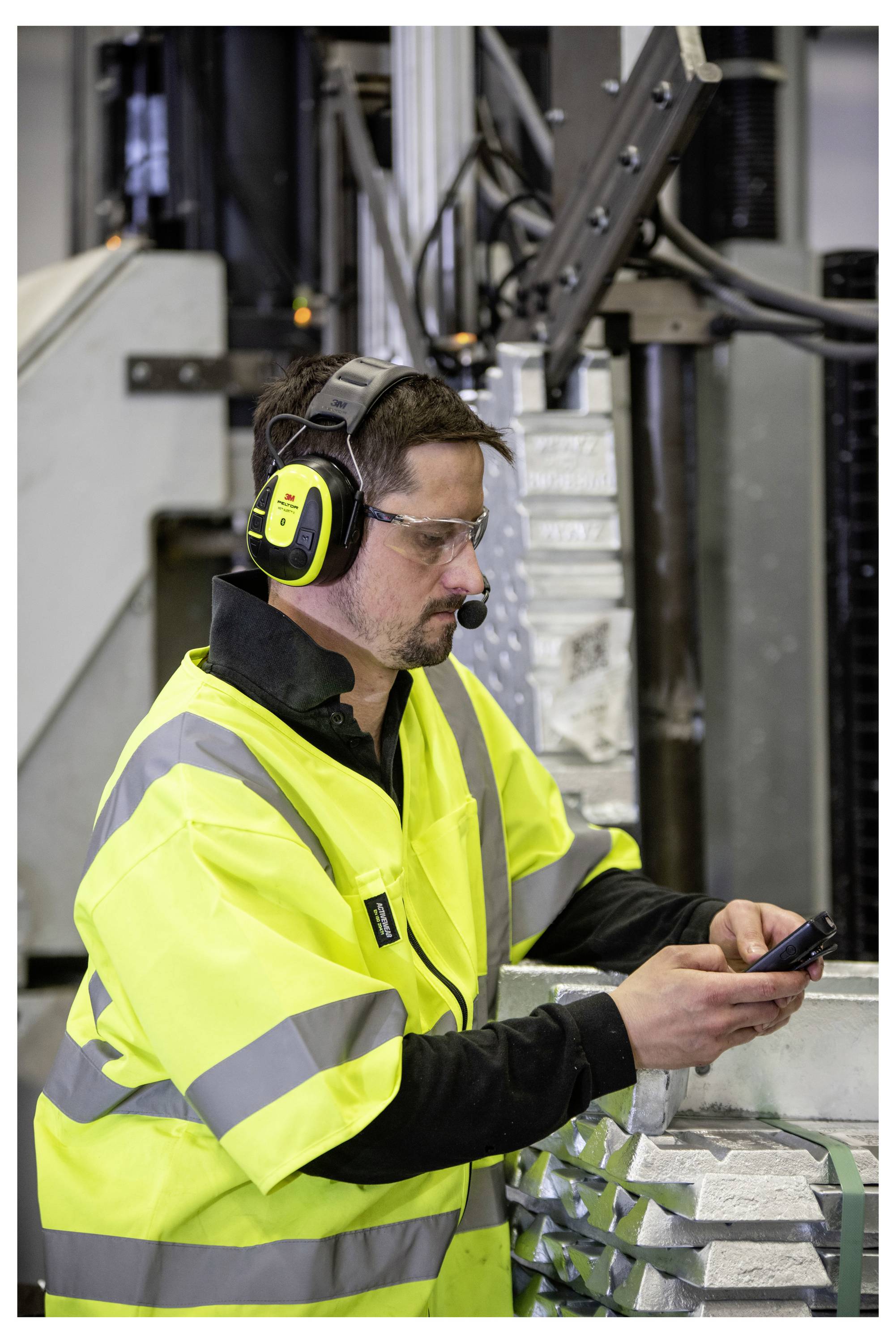En person som bär skyddsutrustning och använder ett headset kontrollerar en enhet i en industriell miljö, vilket tyder på kommunikation eller övervakning av utrustning.