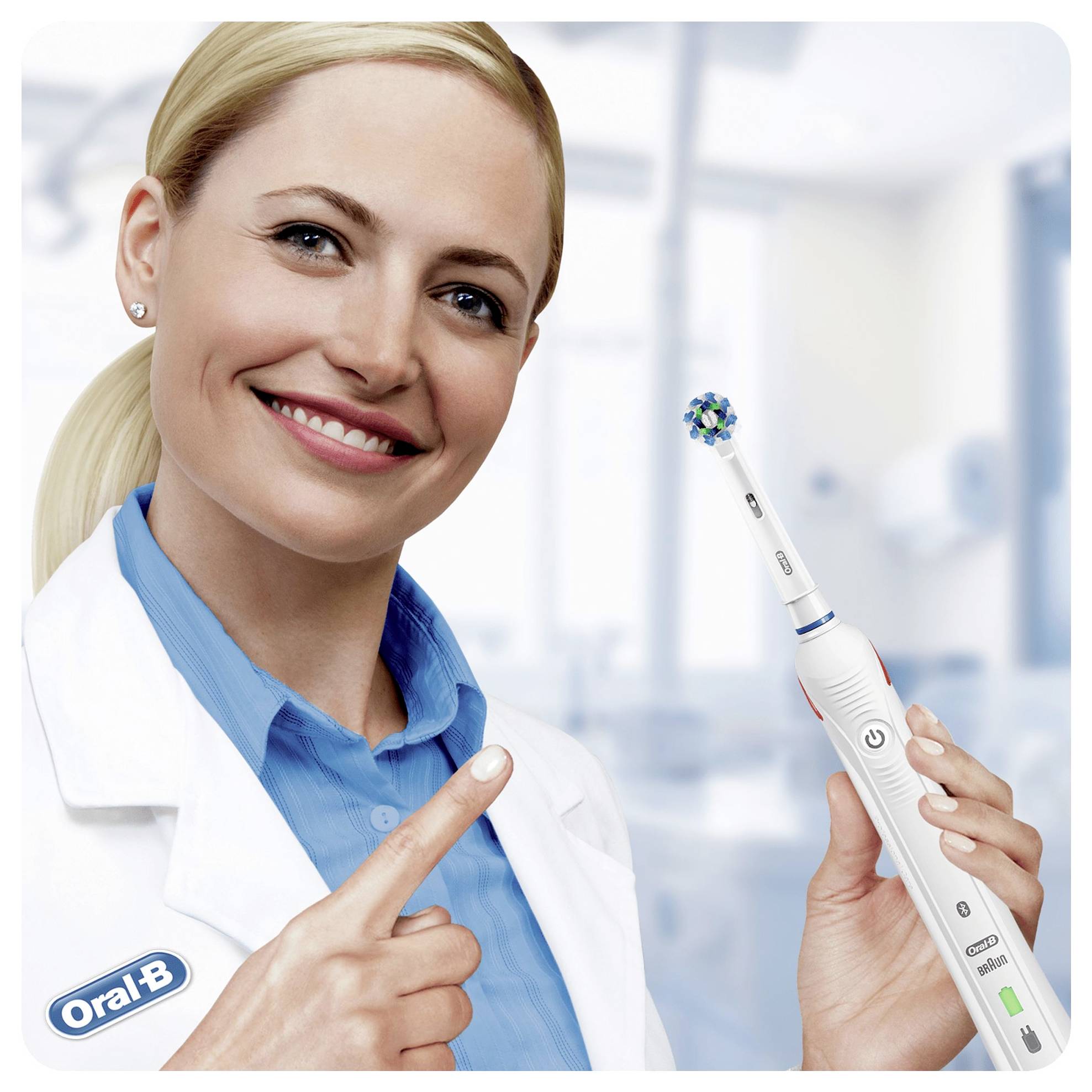 En person i en laboratorierock ler och håller i en Oral-B elektrisk tandborste, och pekar på dess borst, vilket antyder en demonstration eller rekommendation.