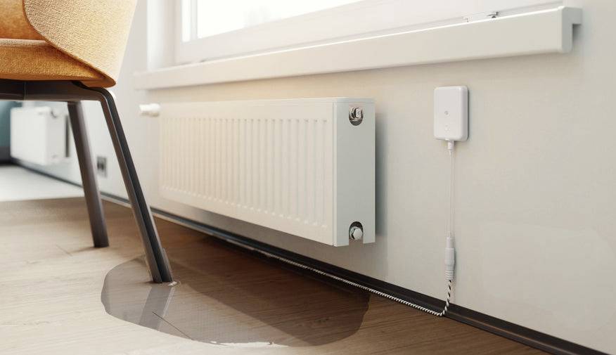 En liten vattenpöl på ett trägolv indikerar en läcka i radiatorn; en vit sensor är fäst i närheten för övervakning.