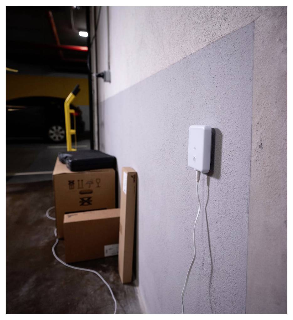 Lådor och en svart resväska är staplade mot en vägg i ett parkeringsgarage, med en enhet monterad på väggen ansluten genom en vit kabel.