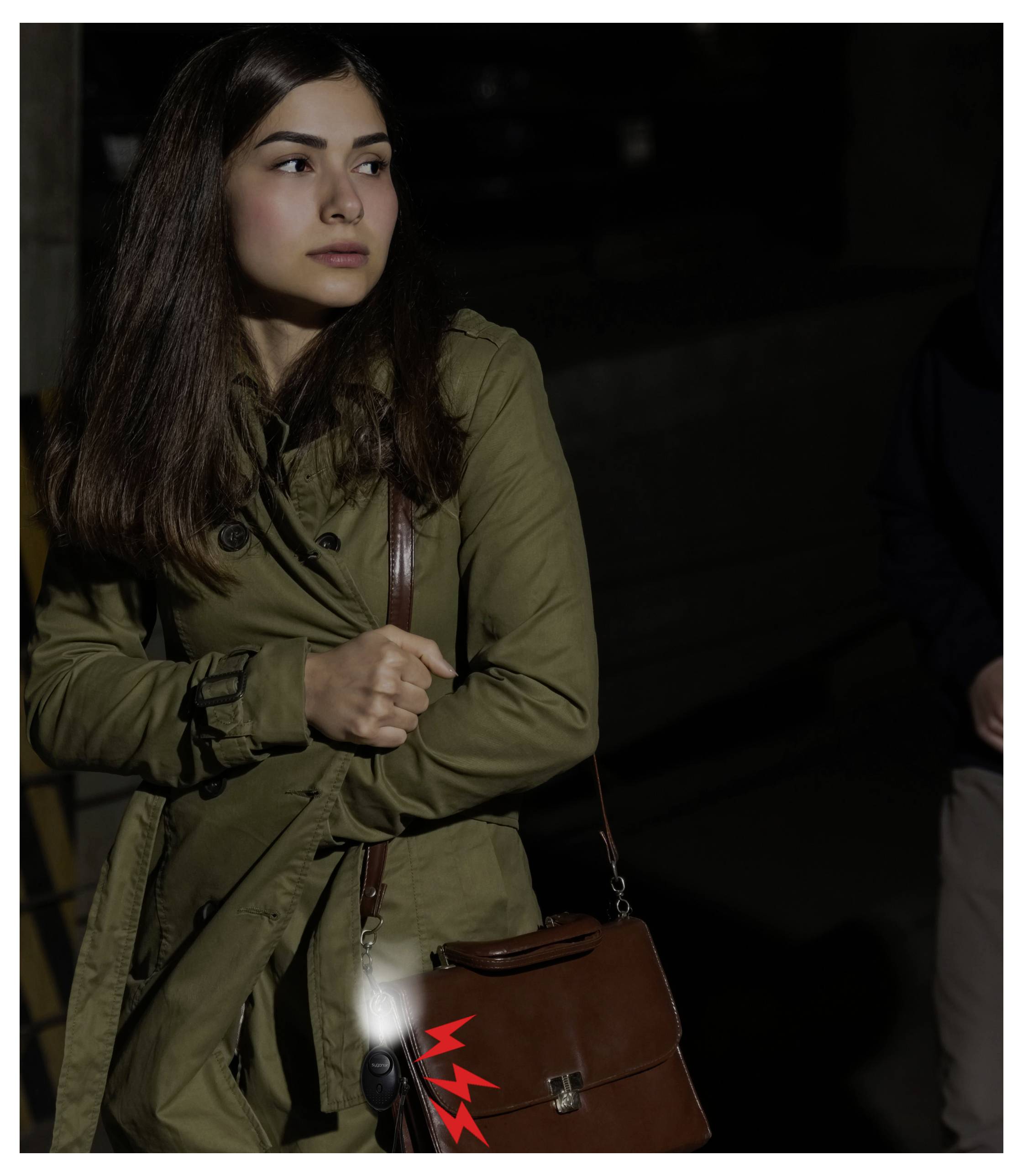 En kvinna i en olivfärgad trench coat håller en brun axelväska, står i svagt ljus och blickar åt sidan.
