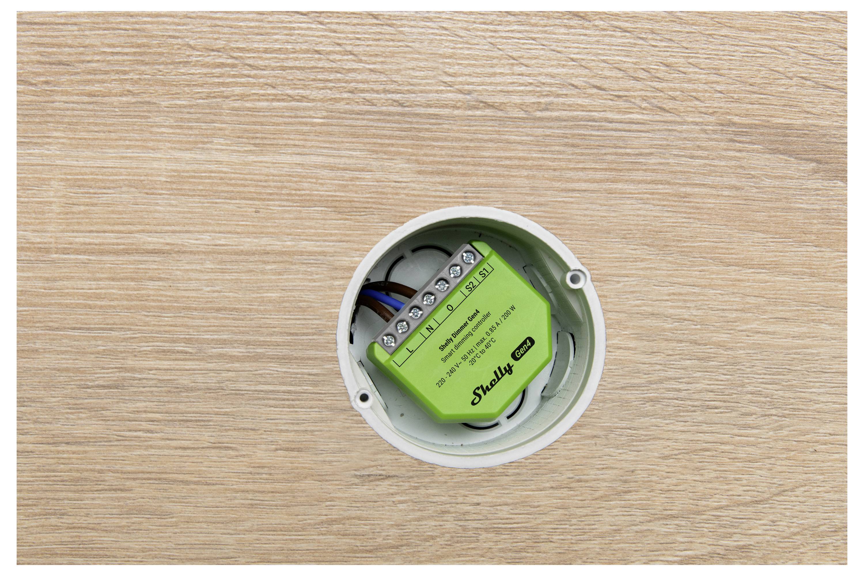 En grön elektrisk enhet är installerad i ett cirkulärt vägguttag. Den är märkt "Shelly 1". Väggen omkring den har en ljus trästruktur.
