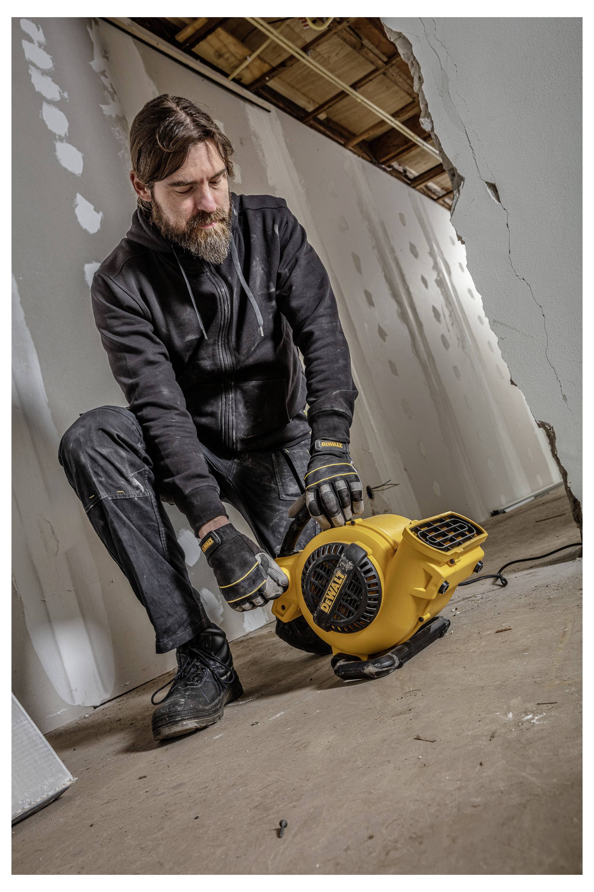 En skäggig man i arbetskläder justerar en gul golvblåsare eller fläkt inuti ett rum som är under renovering med exponerade takbalkar och ofärdiga väggar.