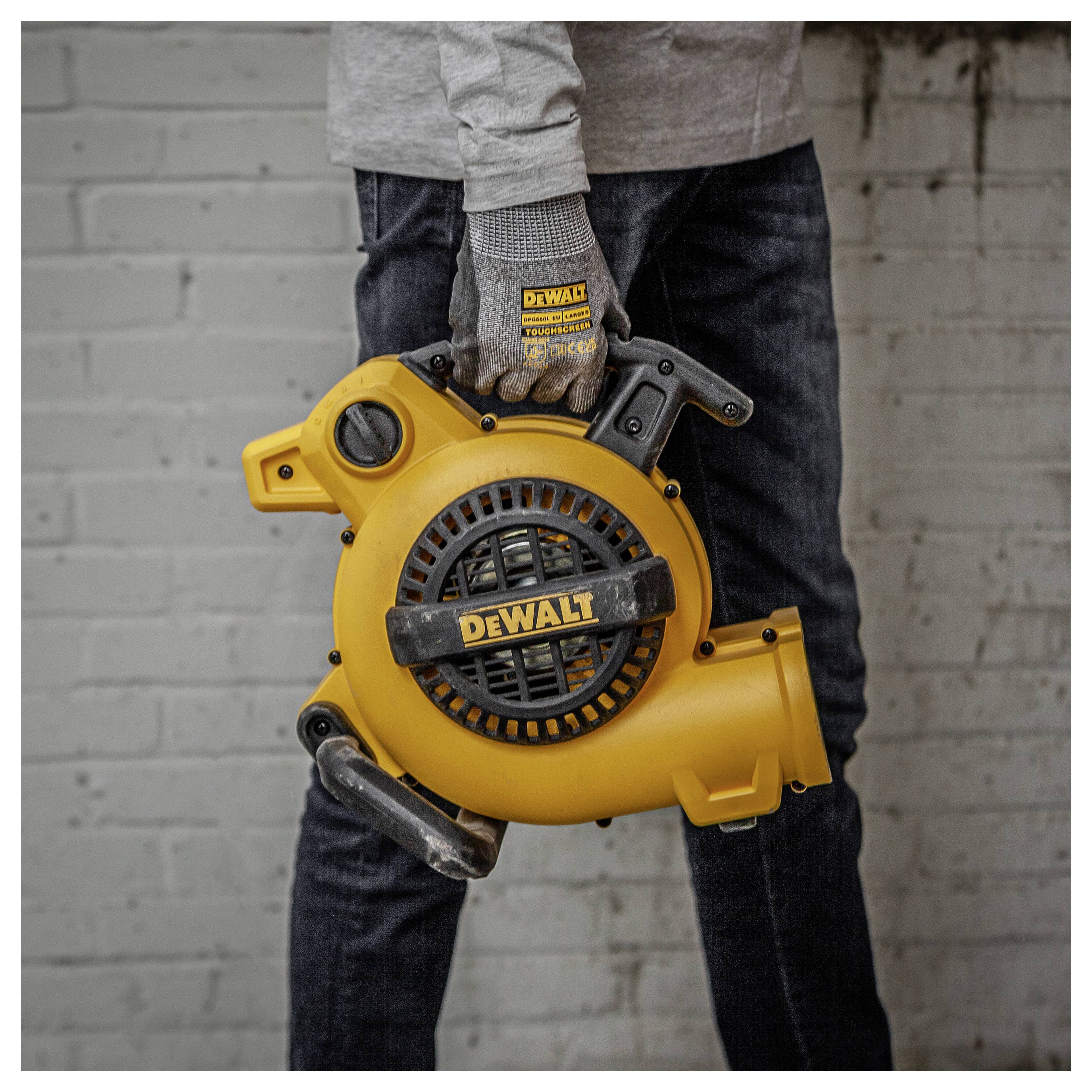 Person som håller en gul DEWALT bärbar blåsare/dammsugare i handtaget mot en grå tegelvägga.