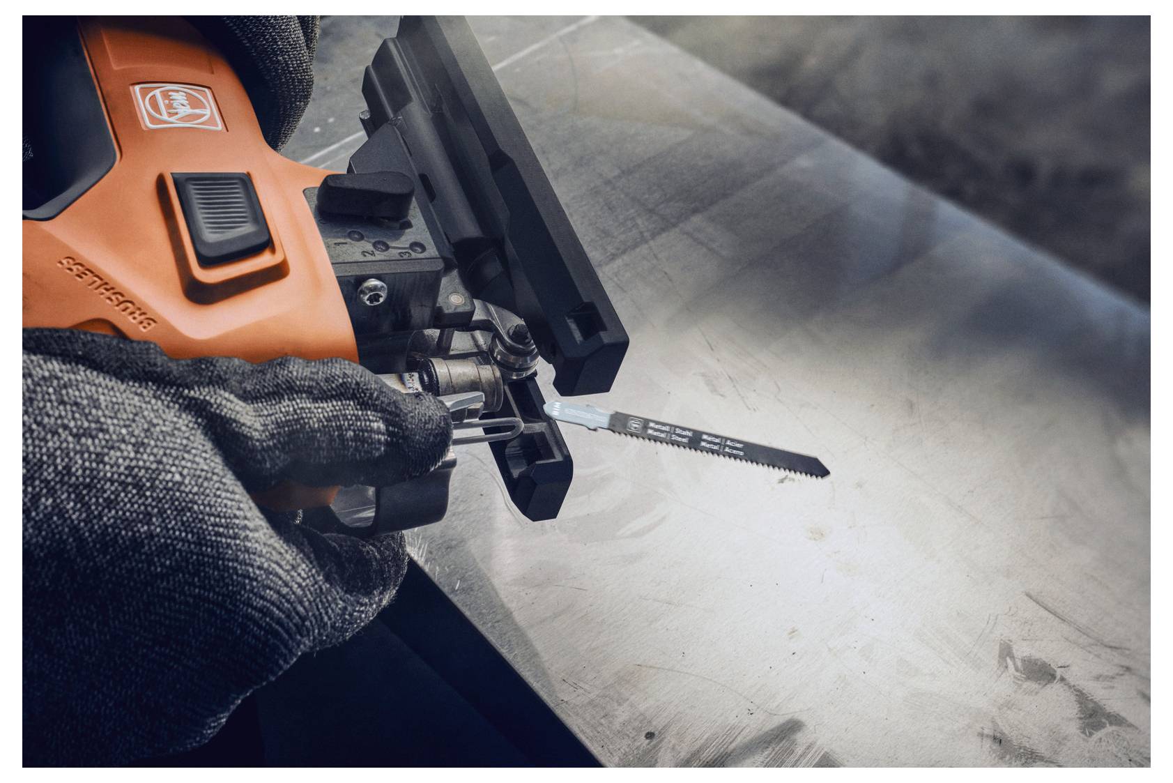 En person som bär handskar använder ett orange och svart elverktyg för att skära genom ett metallstycke på en arbetsbänk. Området är väl belyst.