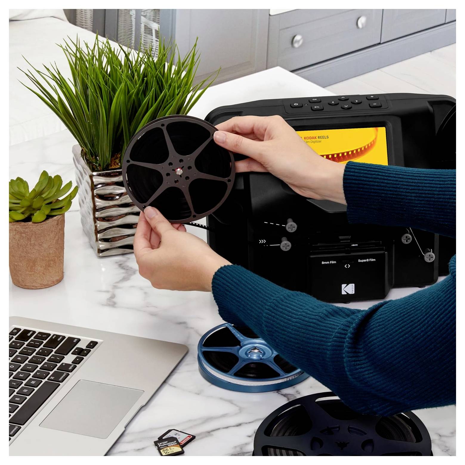 Händer håller en filmspole ovanför en scanningsenhet, med en bärbar dator och minneskortar i närheten på en marmoryta.