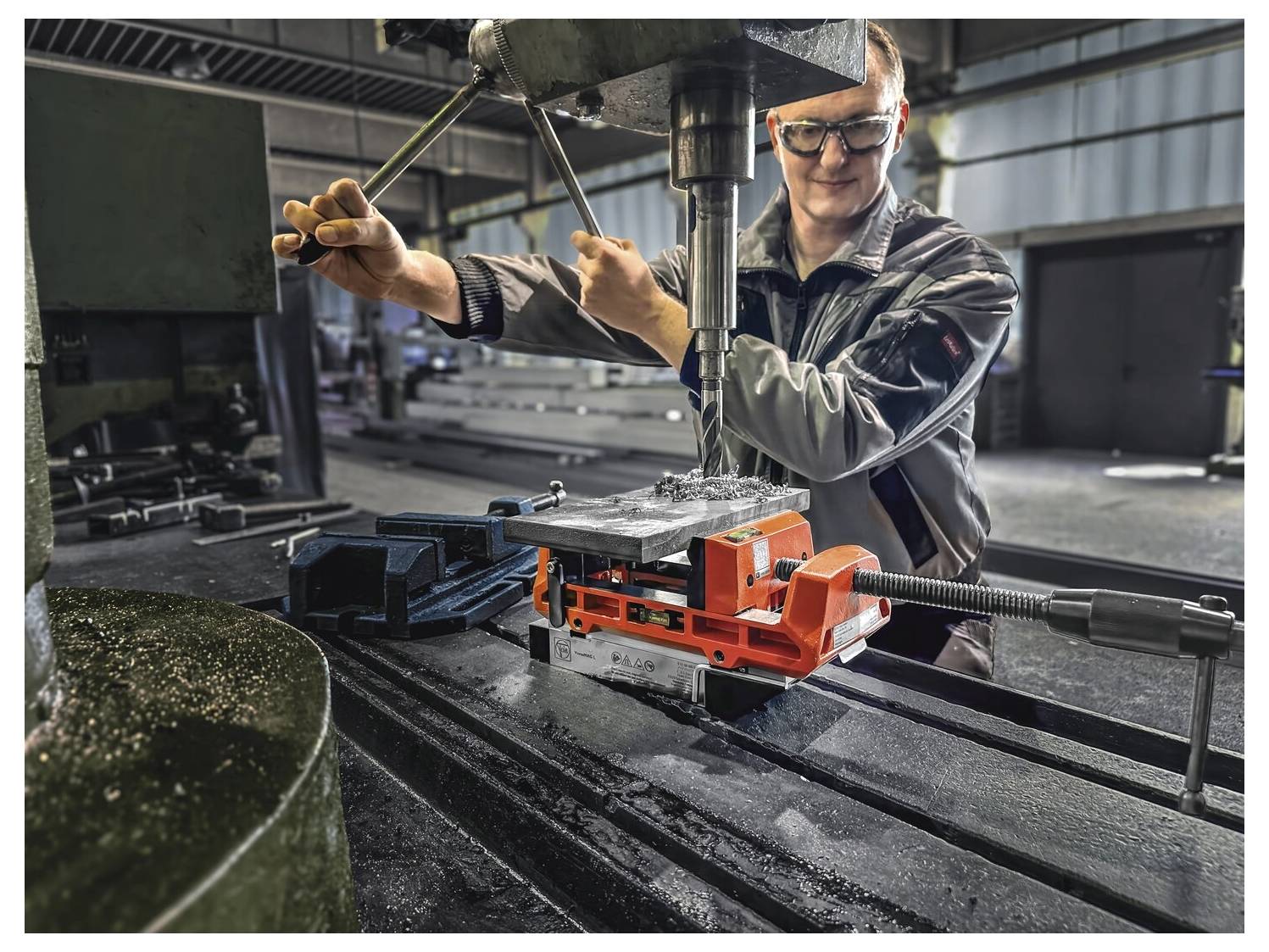 En person använder en industriell borrmaskin i en verkstad och använder ett metallskruvstäd för att säkra ett arbetsstycke för borrning.