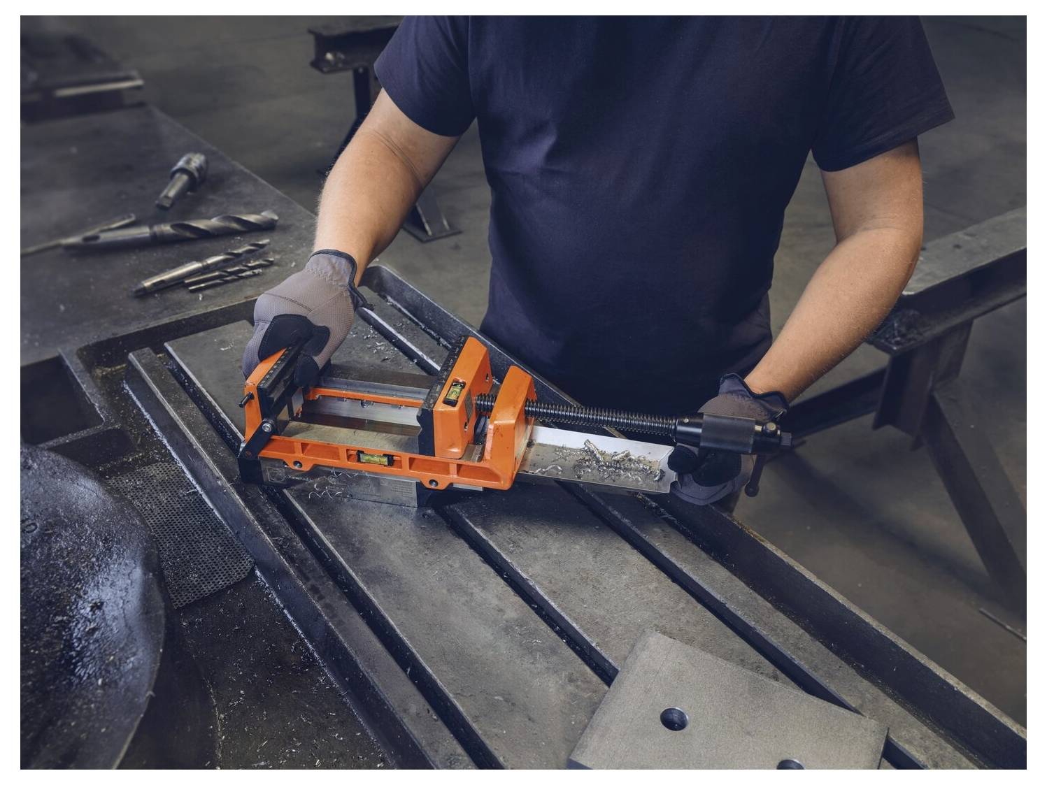 En person använder en orange metallkläm på en arbetsbänk med verktyg i närheten, bärande handskar och en mörk skjorta, i en industriell miljö.
