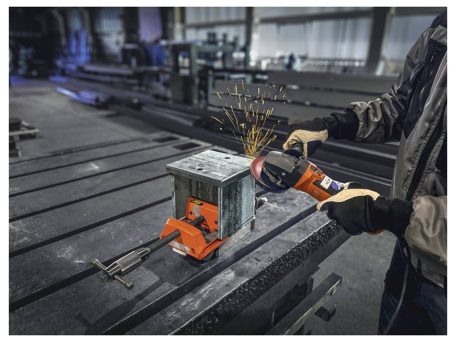 En arbetare sliper en metallåda fastspanad vid ett bord och skapar gnistor. Verkstadsmiljön innehåller industriell utrustning och metallöverflator.