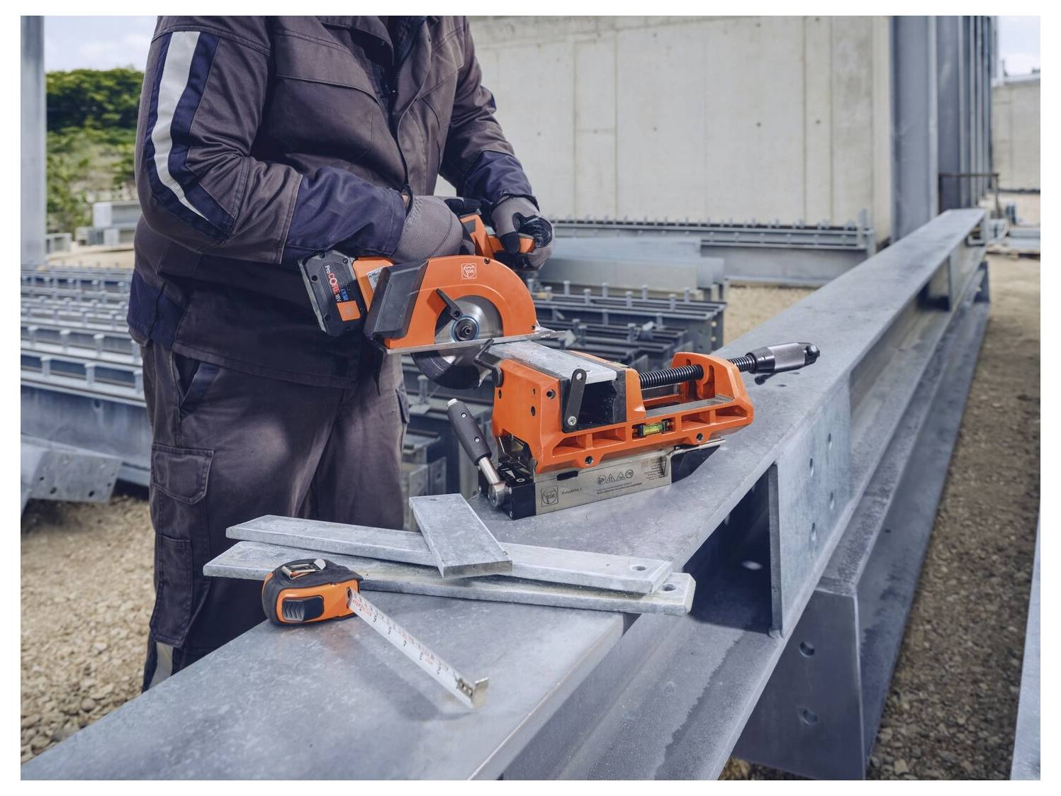 En person använder en orange metallsåg på en stor stålbalk på en byggarbetsplats. Ett måttband och metallvinklar ligger i närheten.