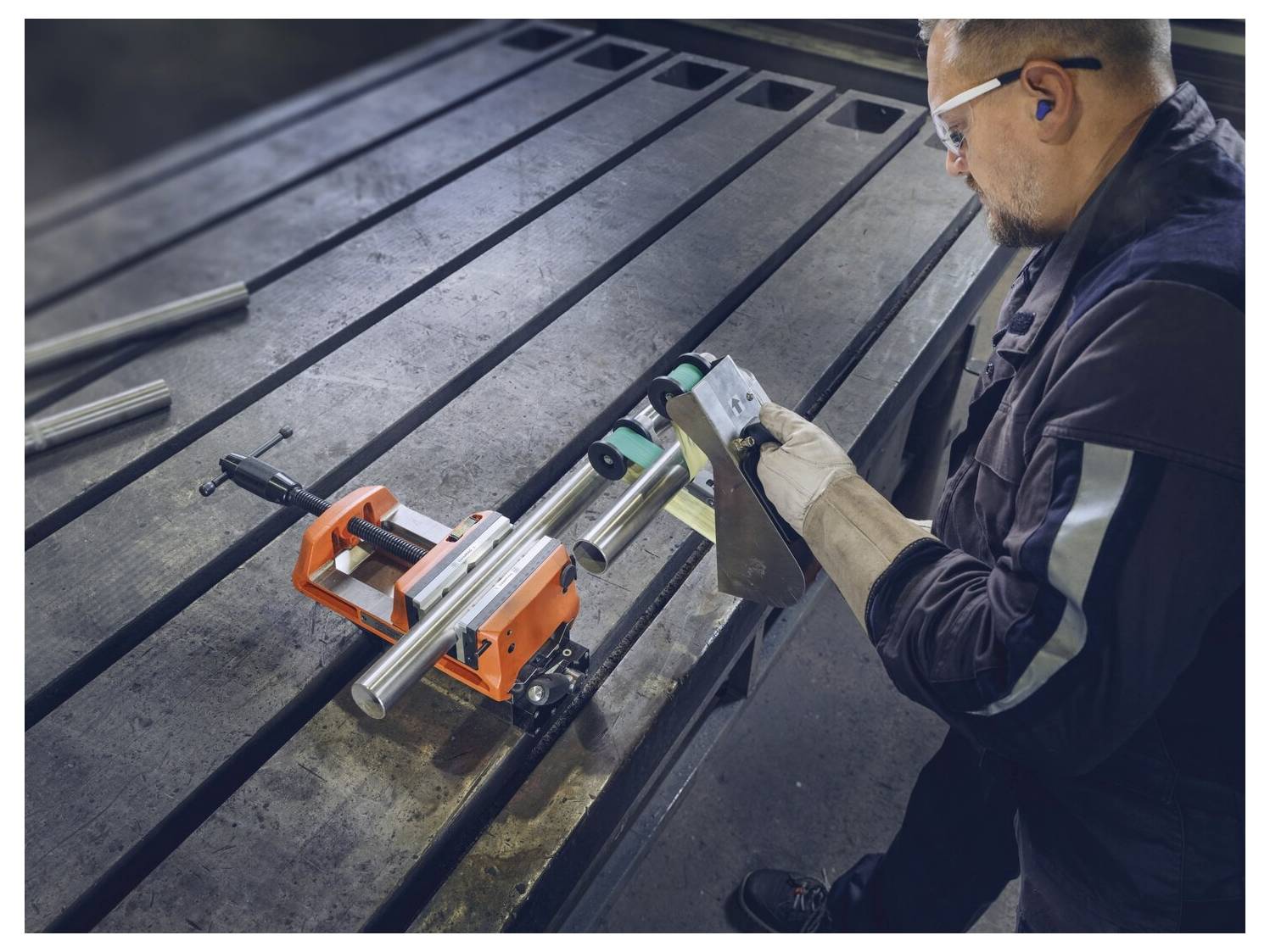 En person i skyddsutrustning manövrerar industriell maskinell utrustning och justerar metallrör med precision på ett stort metallbord i en verkstadsmiljö.