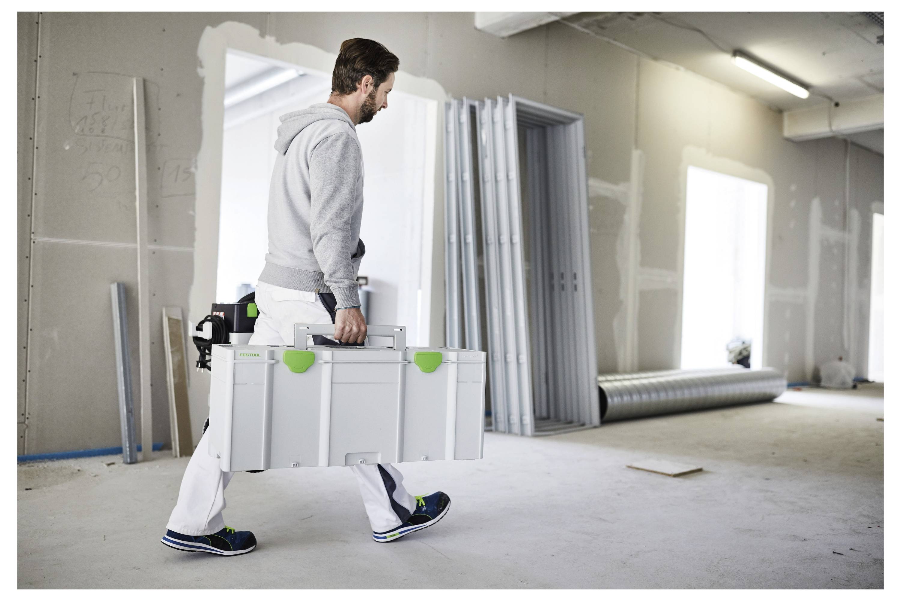 En person i arbetskläder bär en stor, vit verktygsåda i ett rum under renovering. Väggarna är ofärdiga och byggmaterial är synliga.