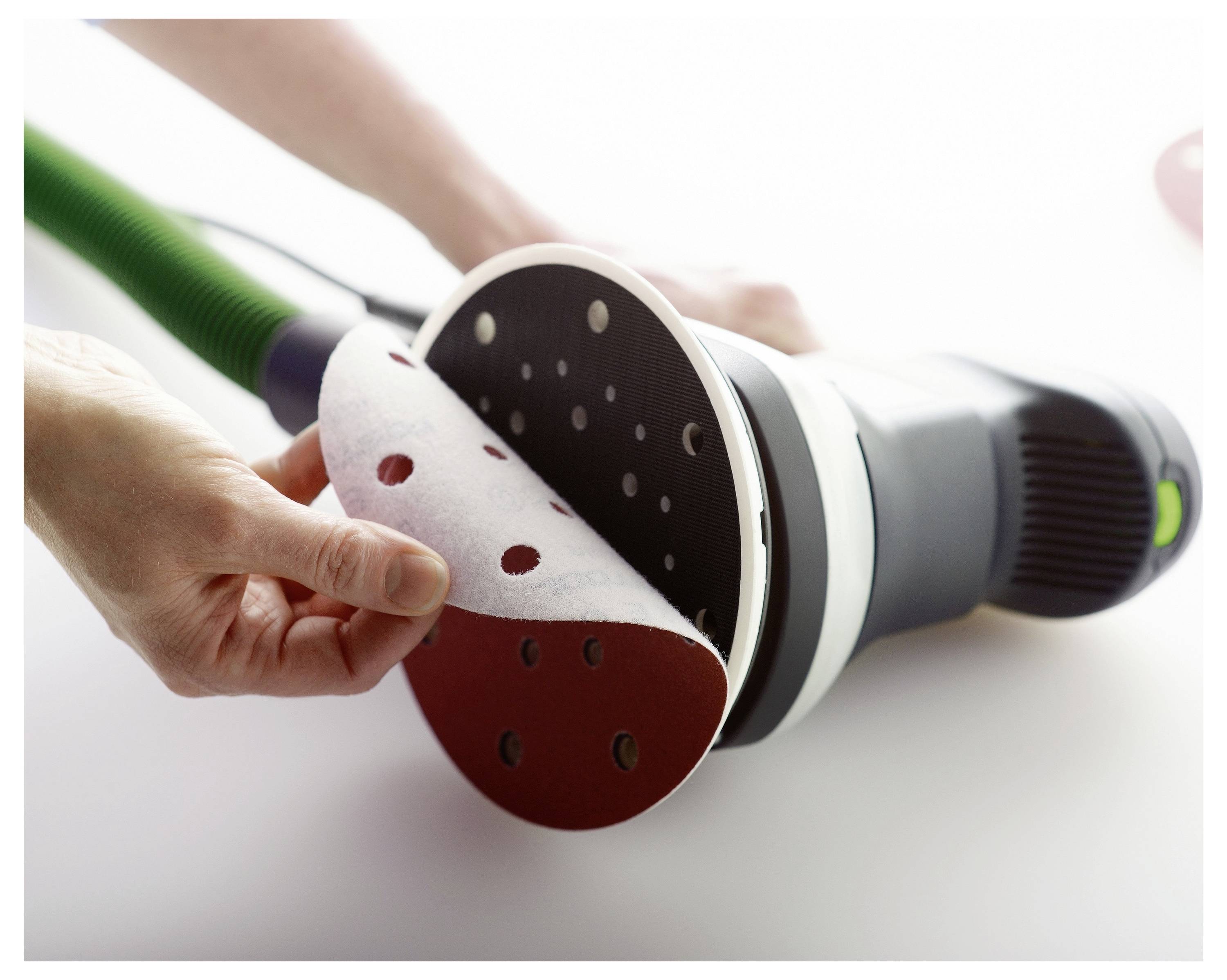 En närbild av händer som byter slippapper på en elslip, som visar justeringen av hål för dammsugning, på en vit yta.