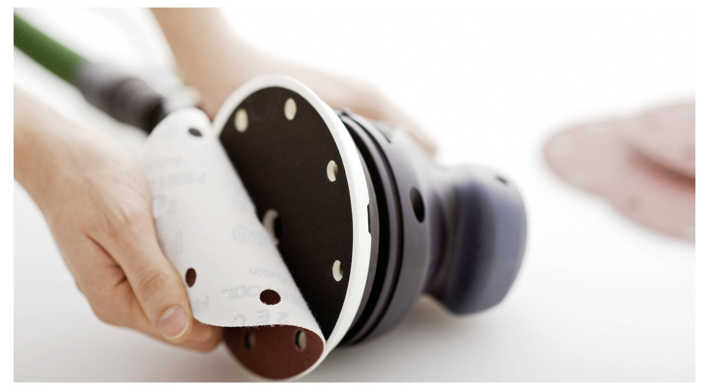 En person använder en handhållen elektrisk sliper på en träyta och fokuserar på att glatta träet. Verktyget är svart med gröna detaljer.