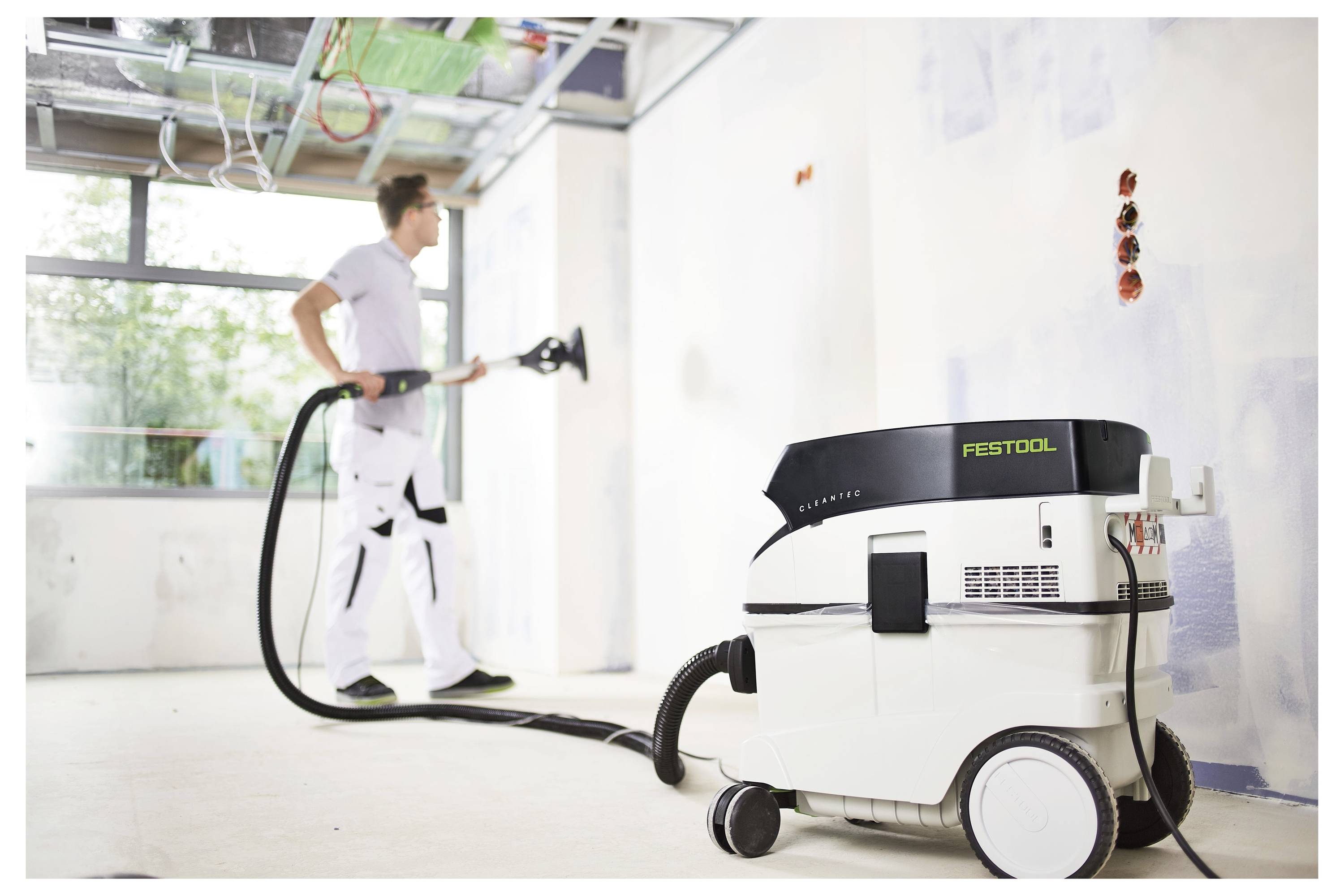En person sliper en vit vägg i ett renoveringsrum, med hjälp av en Festool-maskin ansluten till en dammsugare, med stora fönster i bakgrunden.
