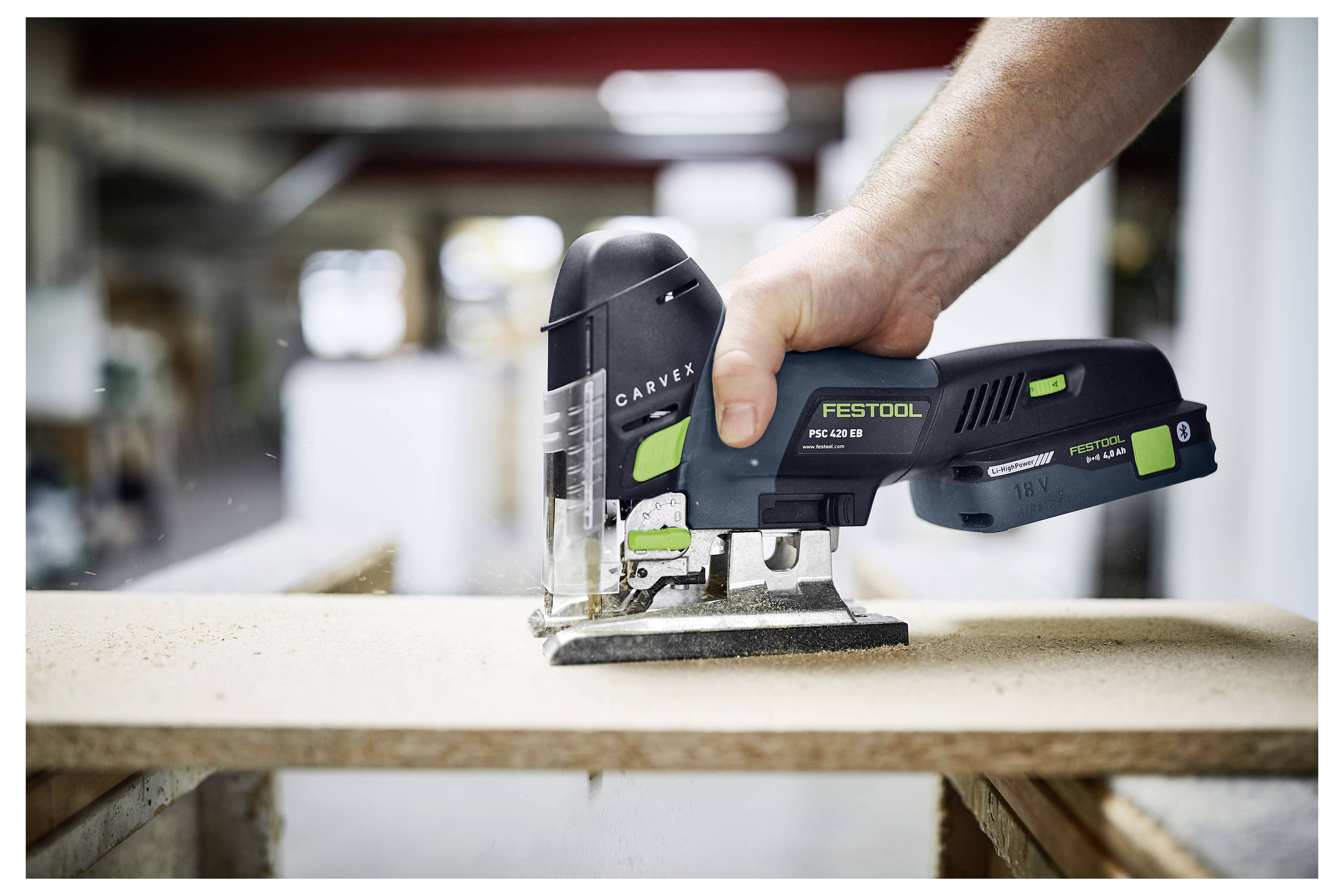 En persons hand som använder en Festool bågfil för att såga genom en trästöd, med sågsågsspån som sprids runt omkring. Bakgrunden visar en suddig verkstadsmiljö.