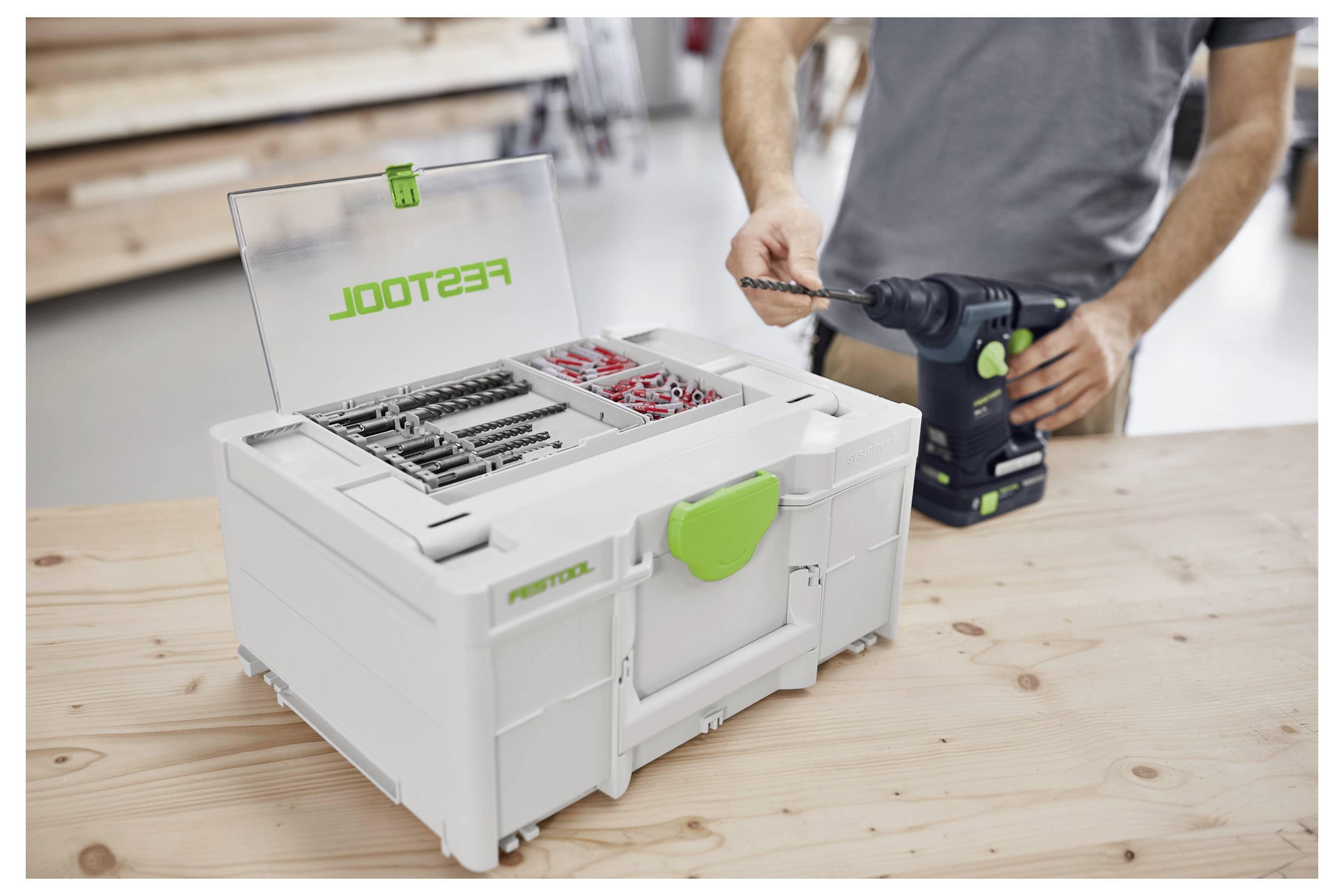 En person håller en elmotordrill bredvid en öppen verktygslåda på ett bord. Verktygslådan innehåller olika borrbitar och är märkt 'Festool'.