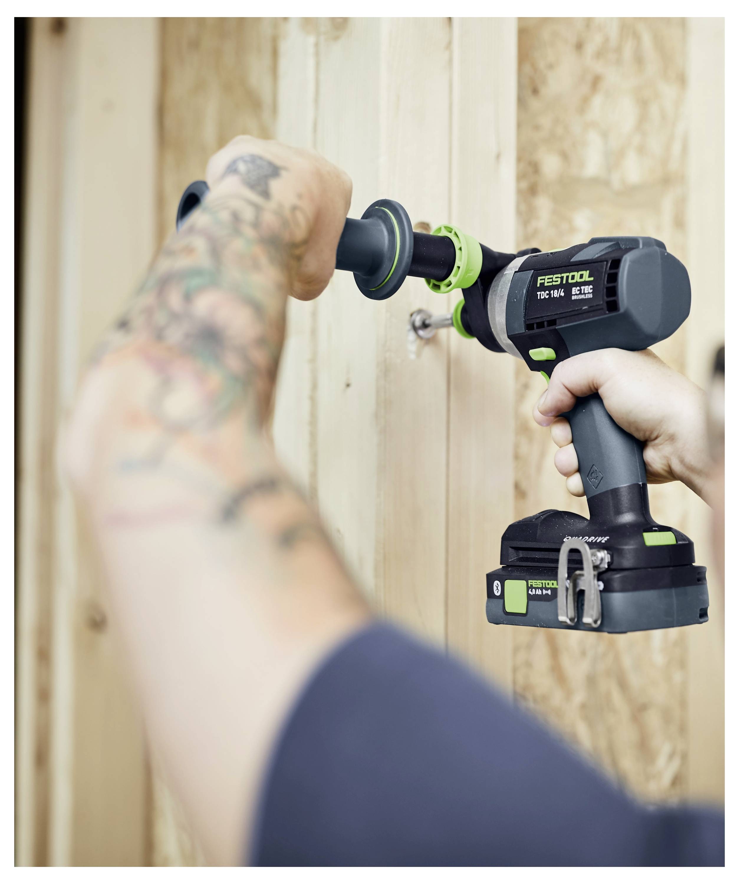 En person som använder en elborr med en tatuerad arm fäster en skruv på en träyta och fokuserar på precision och grepp.
