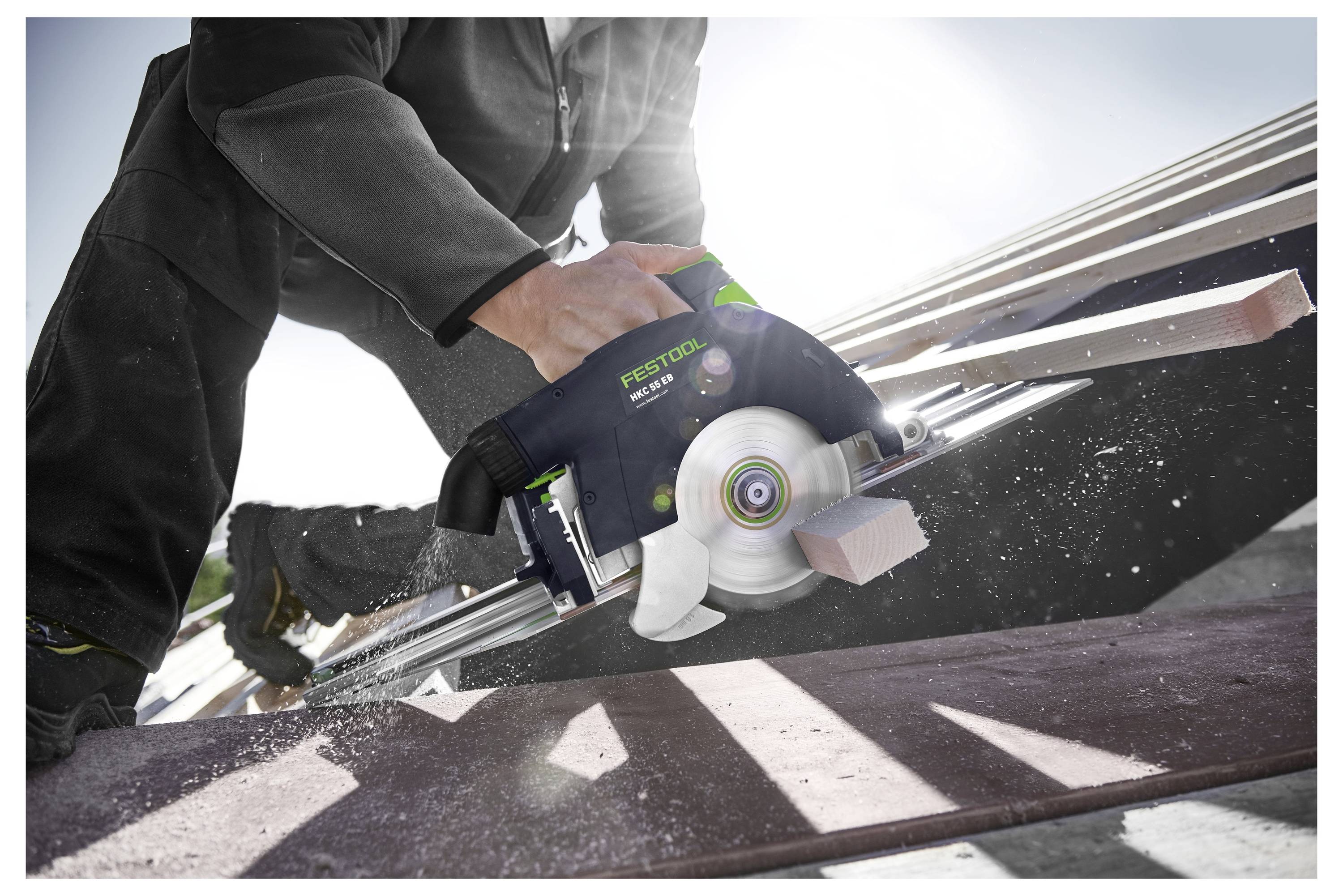 En person använder en Festool-cirkelsåg för att såga genom träbalkar på ett tak, med solljus som skiner i bakgrunden.