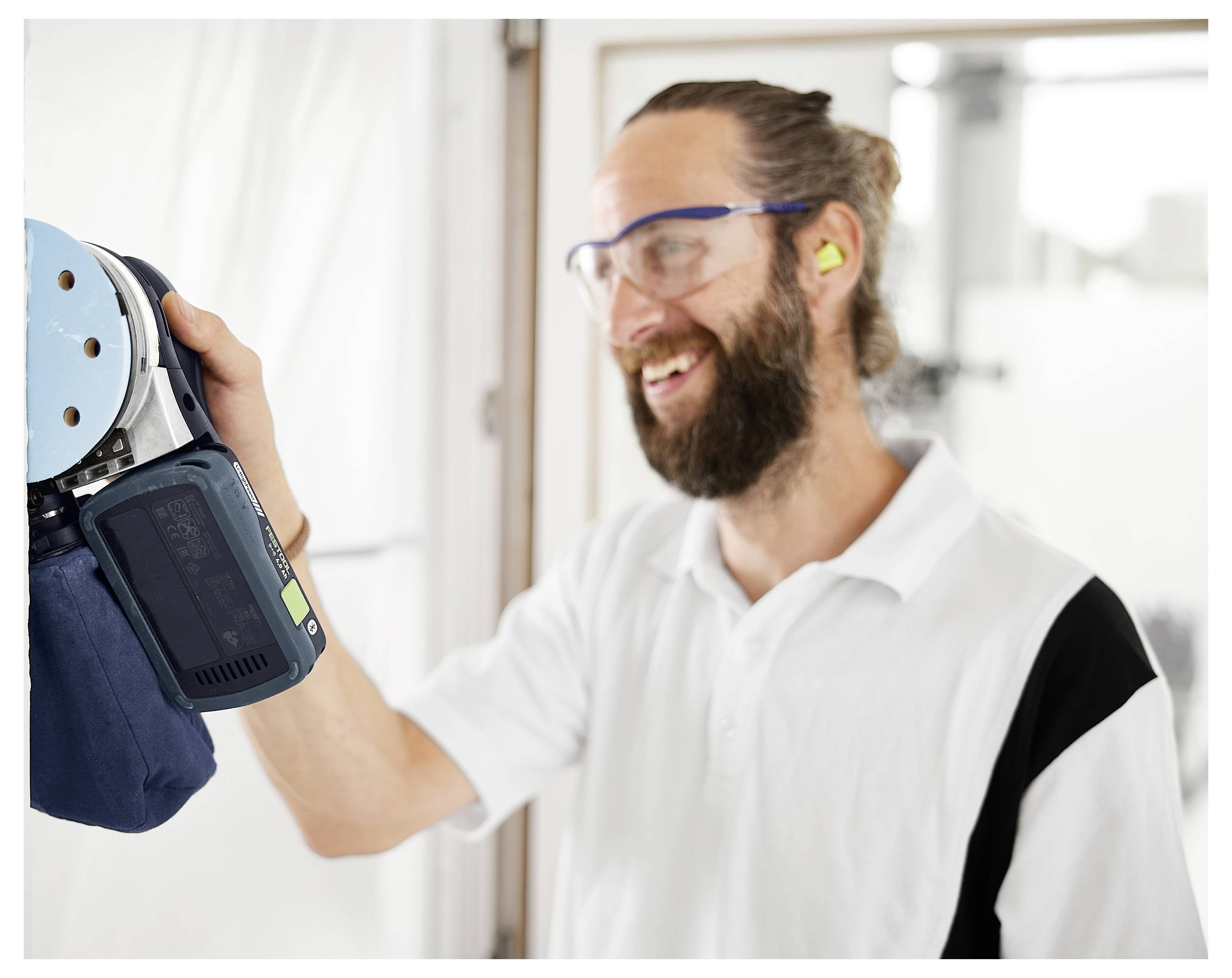 En person som bär säkerhetsglasögon och öronskydd använder en maskin och ler. Miljön verkar vara en verkstad eller ett industriellt utrymme.
