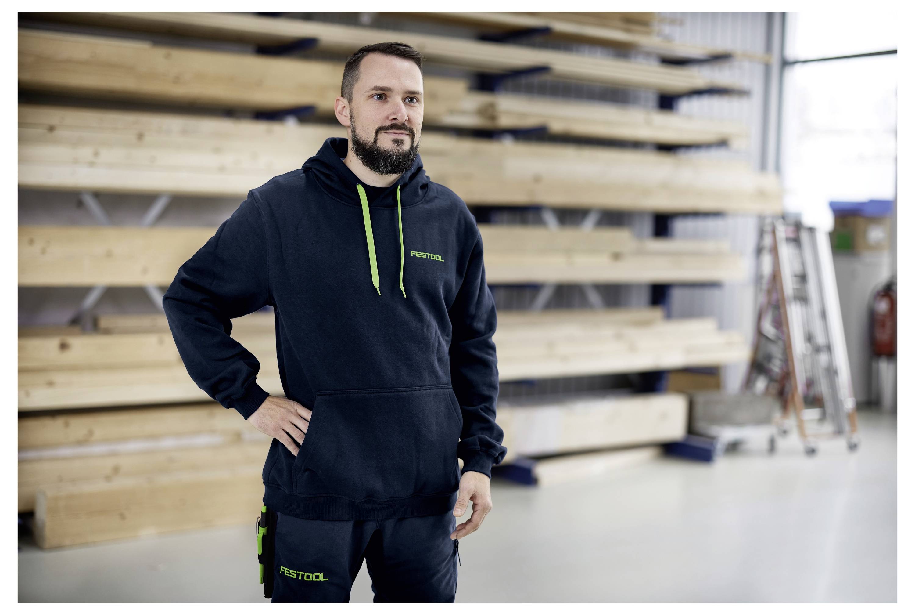 En person i en svart hoodie och arbetskläder står självsäkert i en verkstad, med hyllor och träplankor i bakgrunden.