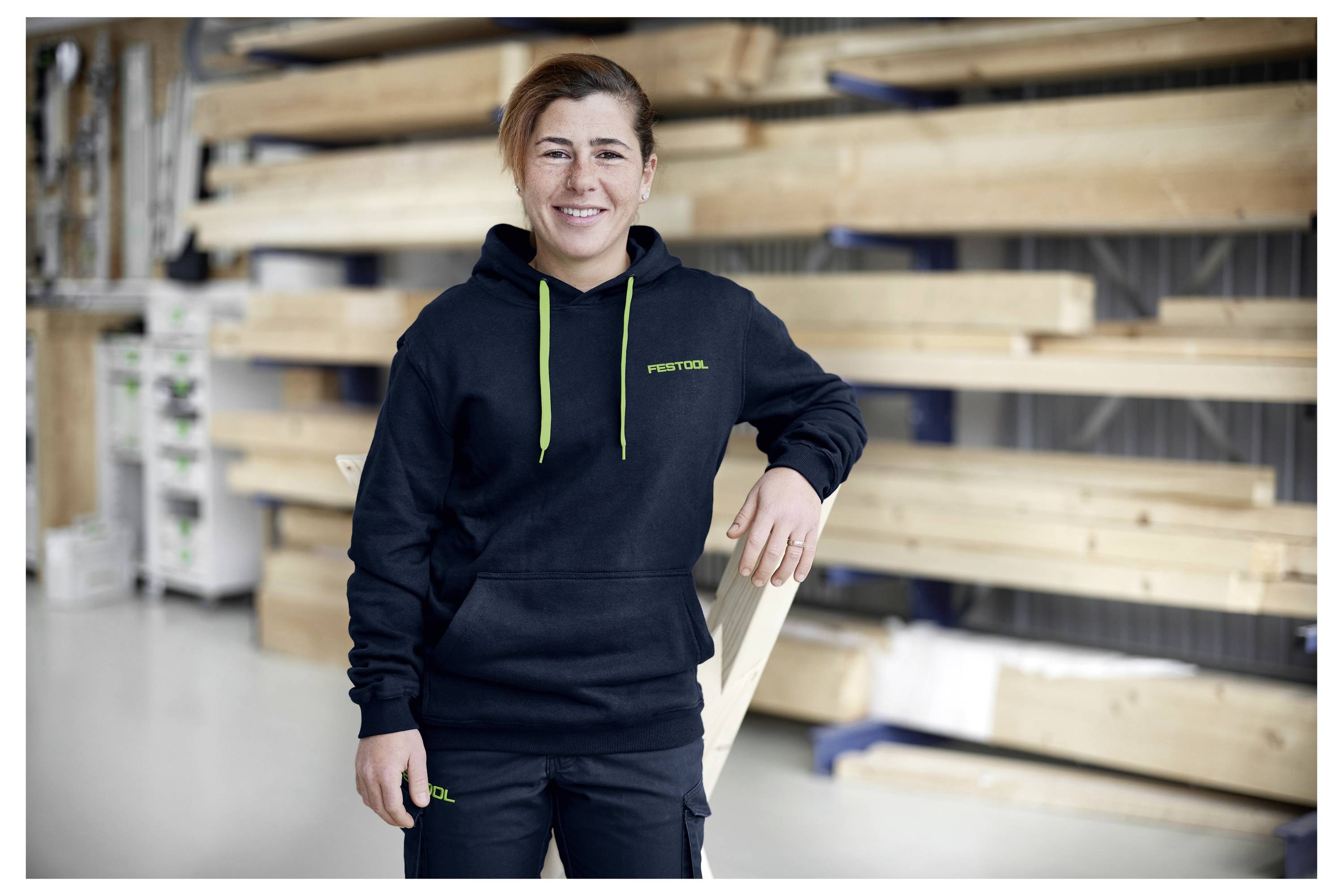 En person i en workshop som bär en svart hoodie med "Festool" på den, som står framför staplade träplankor och ler mot kameran.