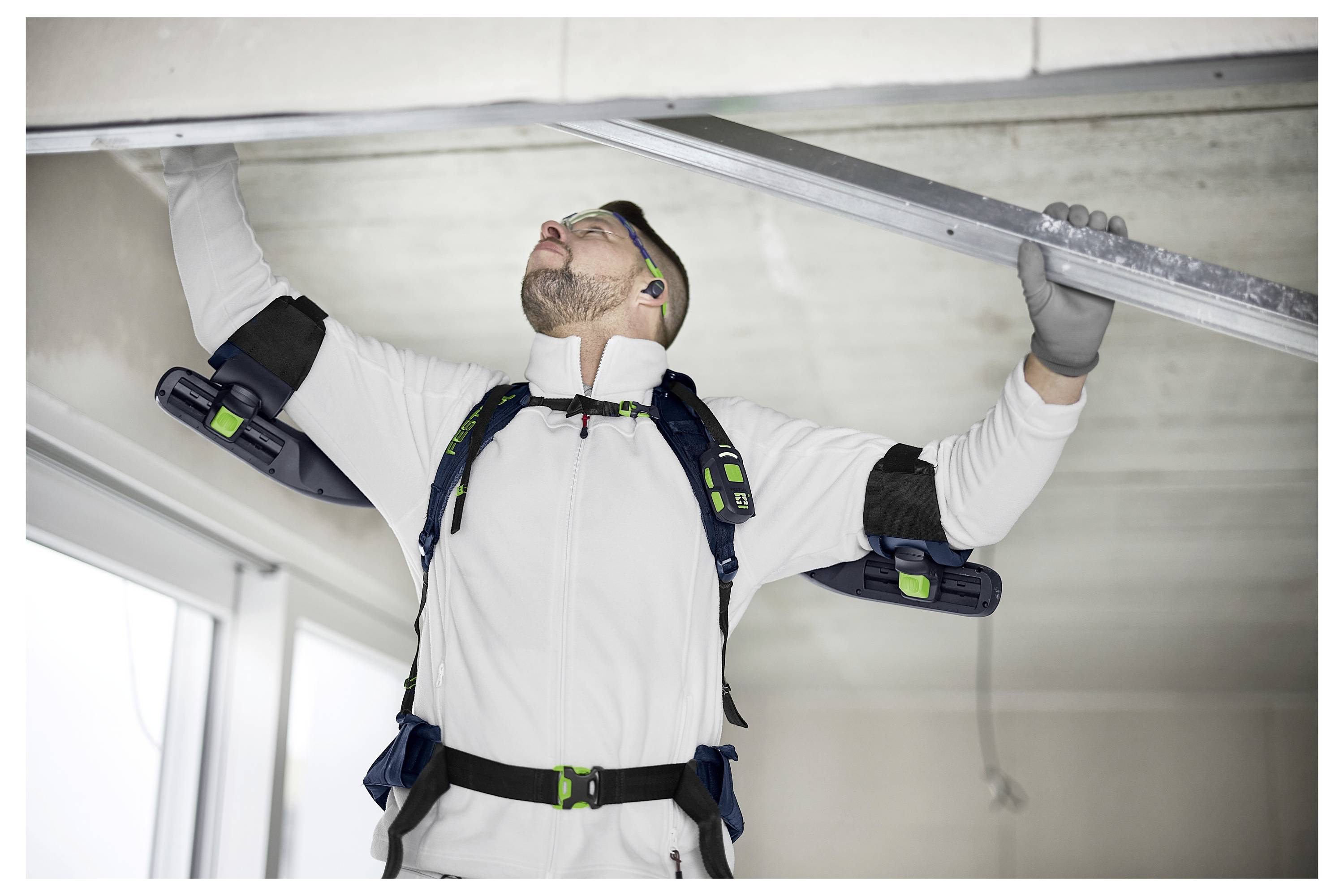 Byggnadsarbetare installerar en metalltak panel, klädd i säkerhetsutrustning och en elektromekanisk exoskelett för stöd i ett ljust rum.