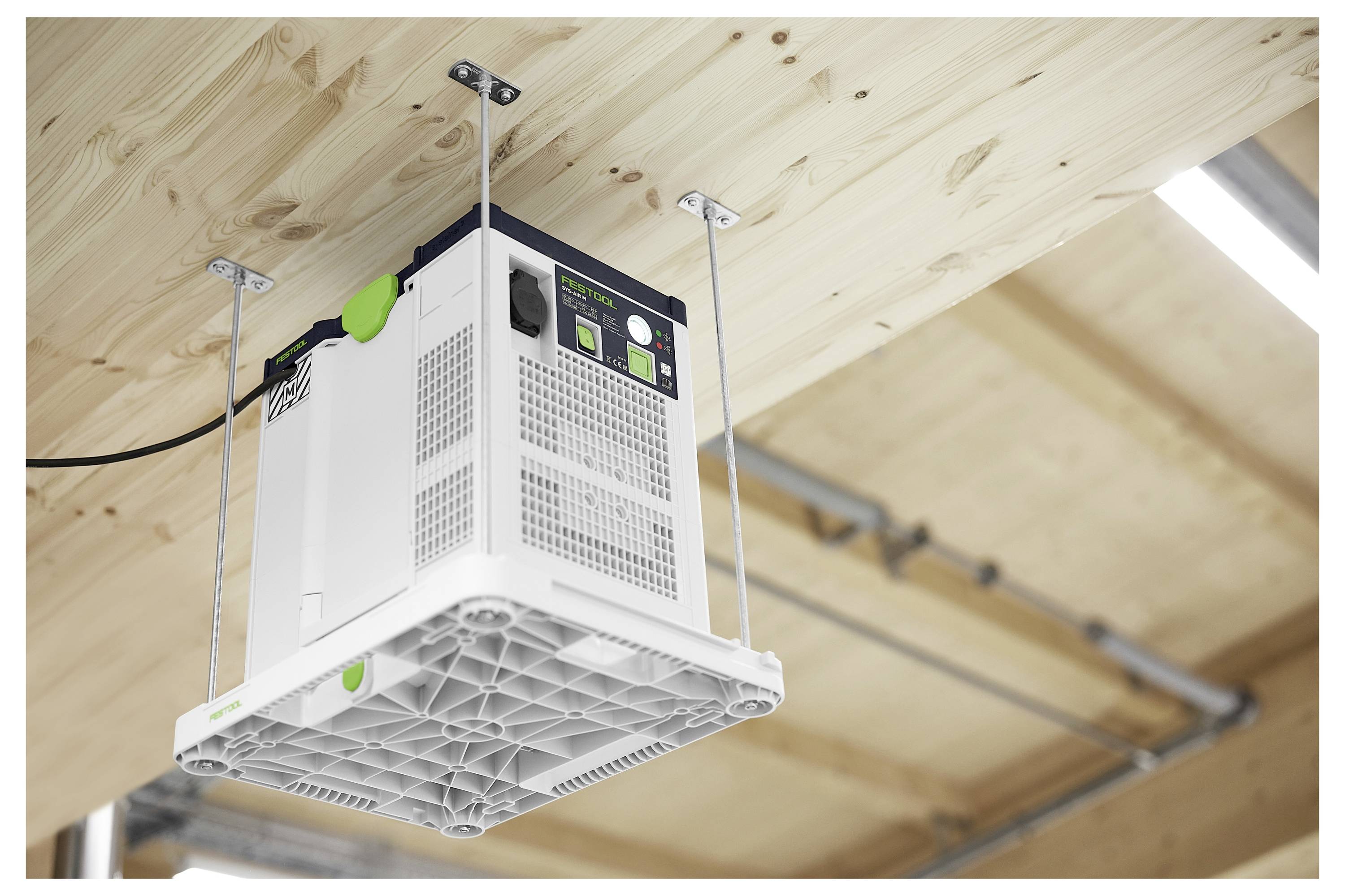 En vit luftfiltreringsenhet är monterad upp-och-ned på ett trägolv med metallkonsolar i en verkstadsmiljö.