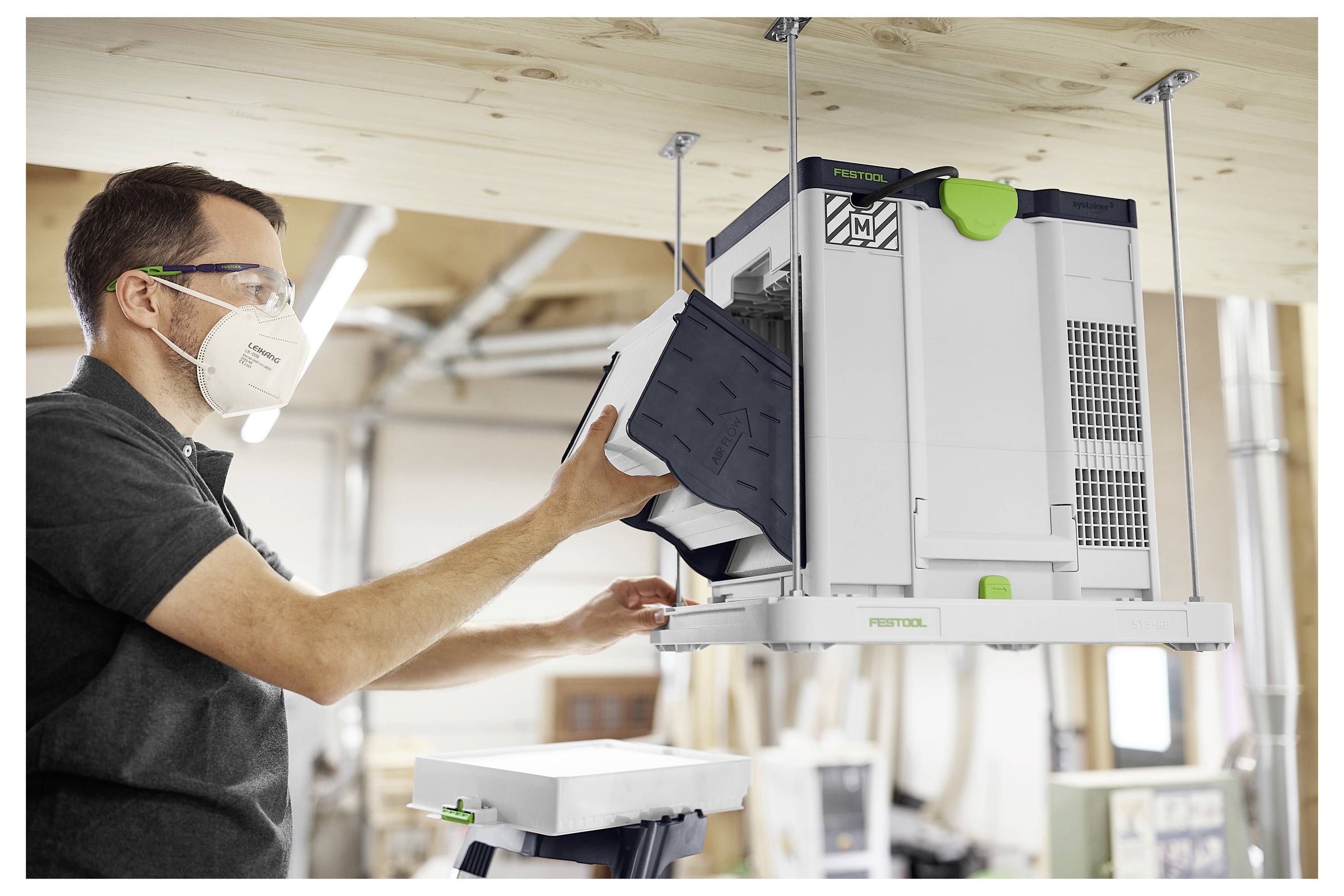 En person installerar ett ventilationssystem på ett trättak i en verkstad, medan han bär skyddsglasögon och en mask för säkerhet.