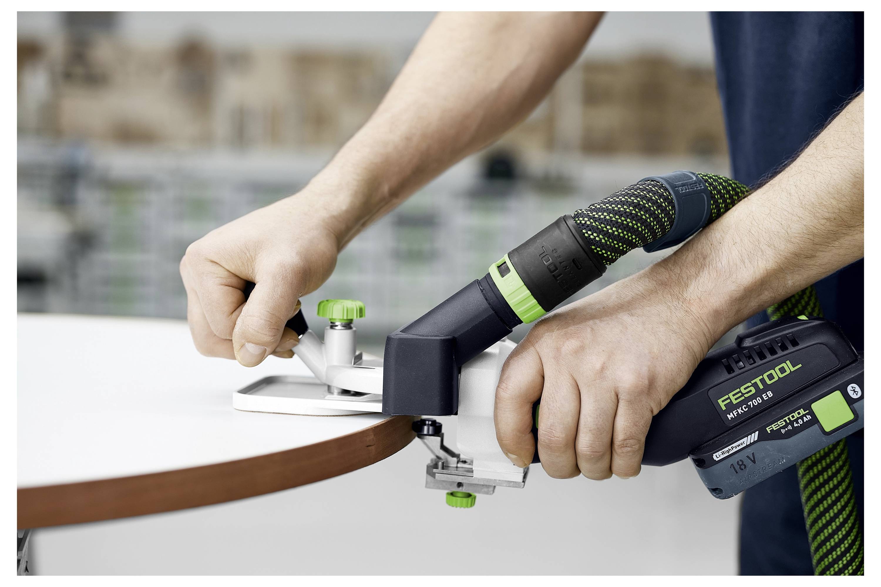 En person använder ett trådlöst kantsliparverktyg för att applicera kantbeslåg på en träbräda. Verktyget är grönt och svart och märkt "Festool".