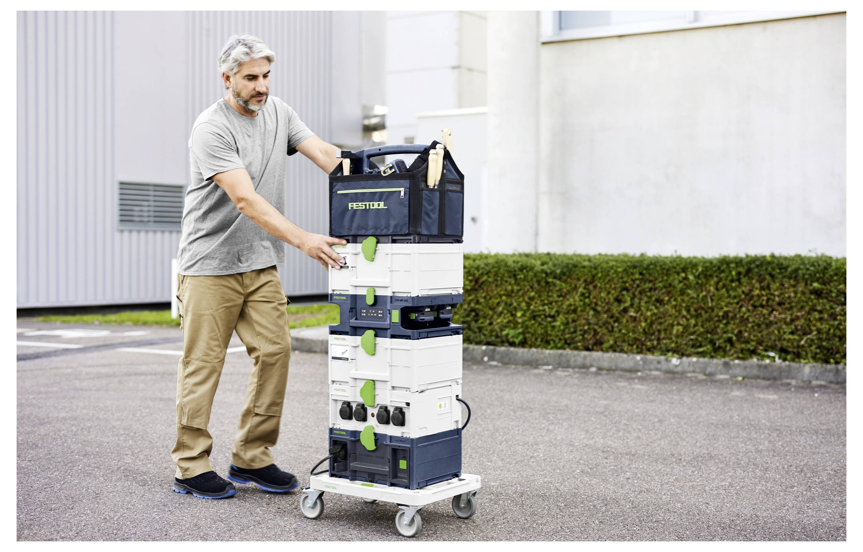 En person skjuter en stapel med Festool-utrustning på en hjälpvagn utomhus. Utrustningen är organiserad i modulära behållare.