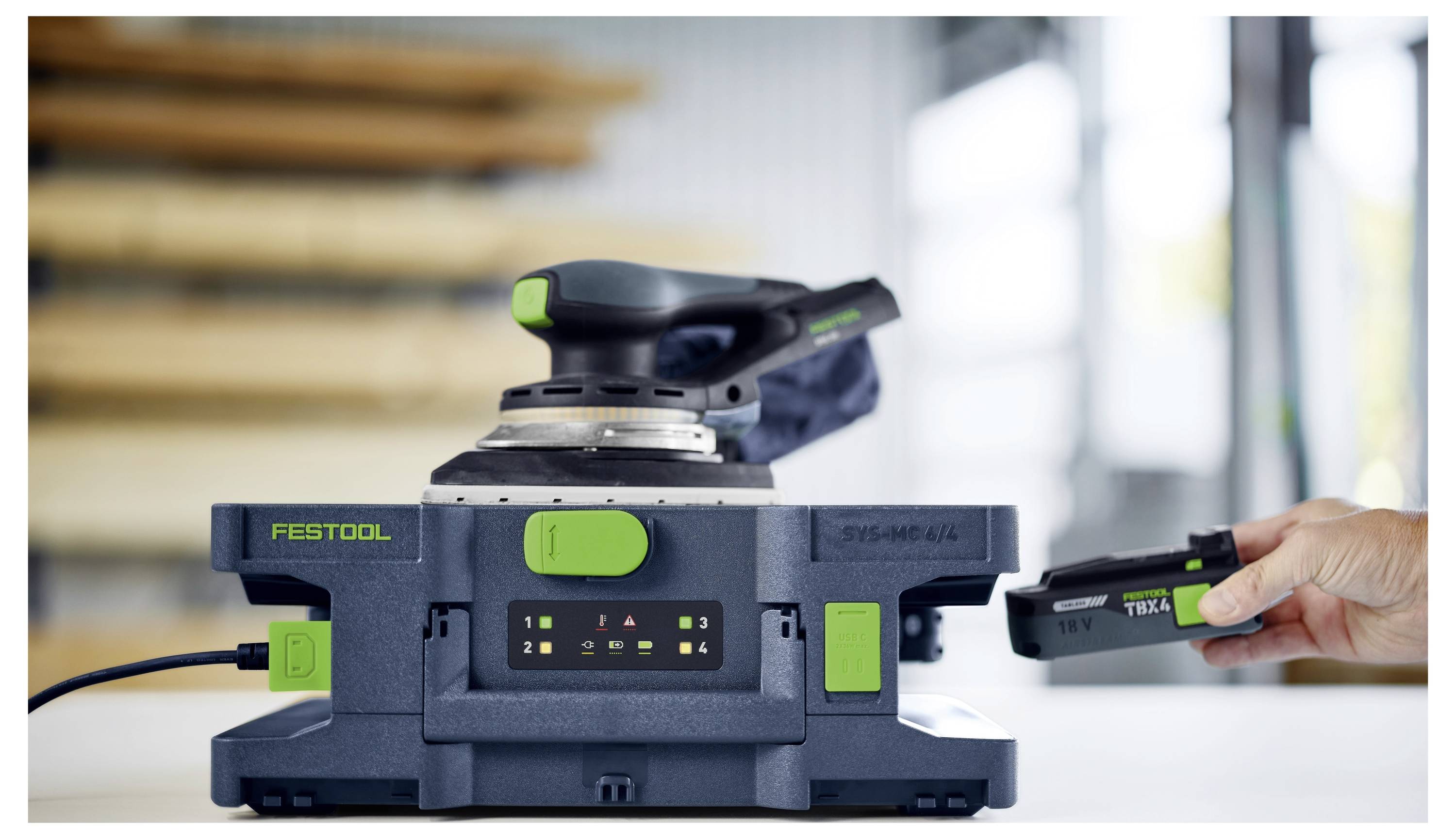 En hand sätter ett batteri i en Festool SYSLITE KAL II arbetslampa, med en trådlös slipmaskin vilandes på en arbetsbänk i bakgrunden.