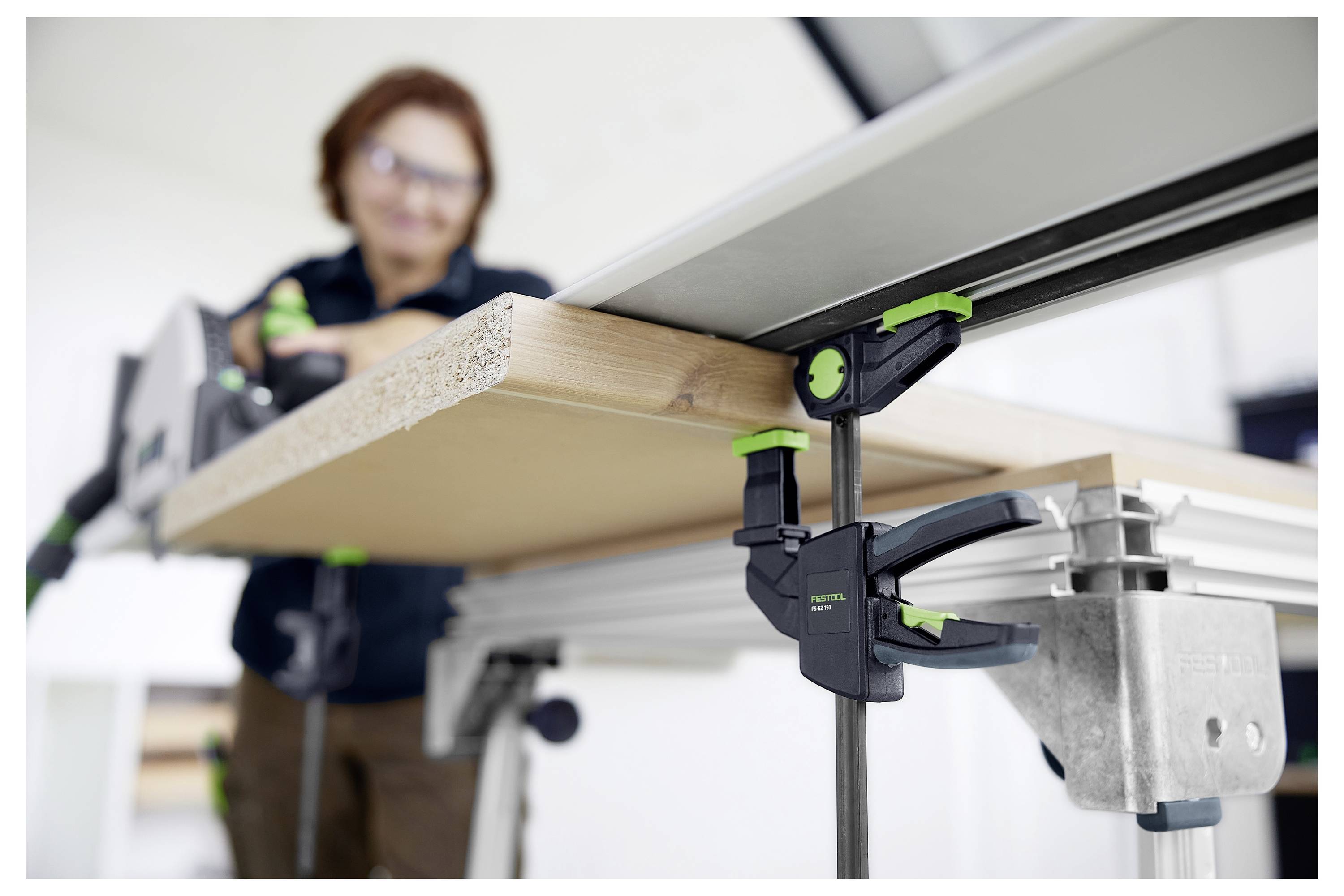 En person använder gröna och svarta skruvspreader för att fästa ett träbräde på en bordssåg, med fokus på precisionsträverk.