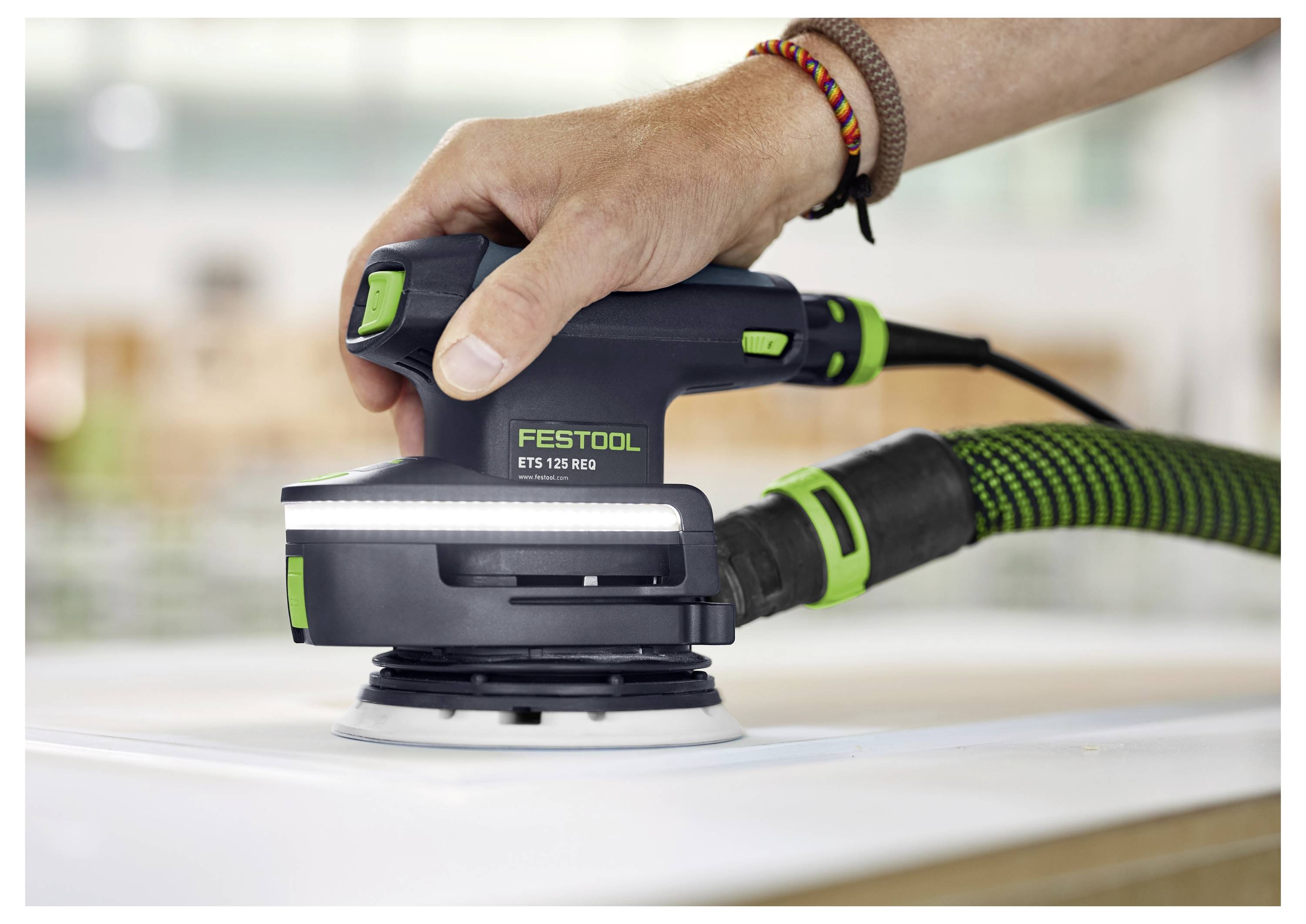 En hand som använder en Festool ETS 125 REQ slipmaskin på en plan yta, ansluten till en dammsugarslang, och demonstrerar dess slipförmåga.