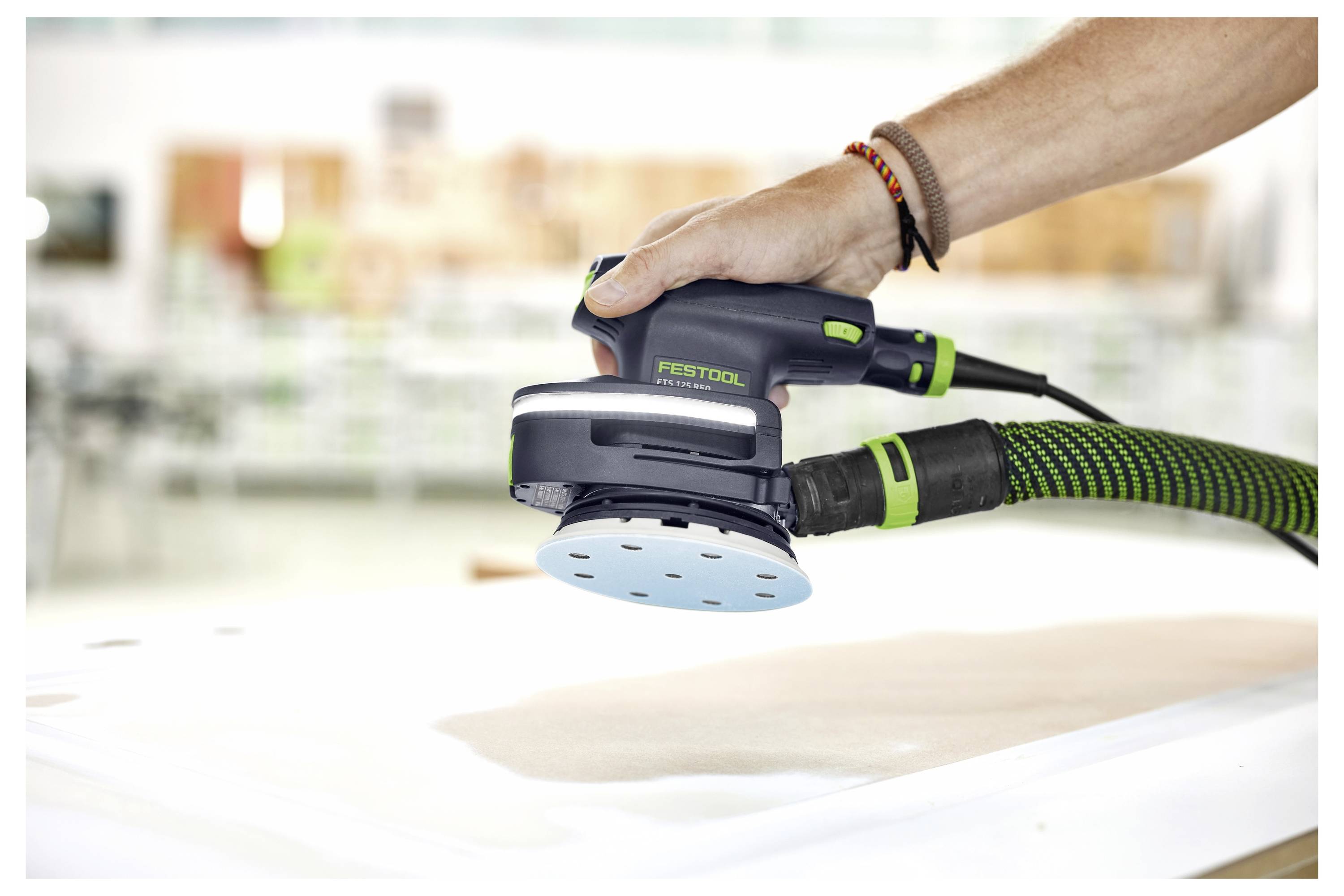 En hand som håller en Festool-elslip som arbetar på en plan trävyra, med en slang ansluten för dammextrahering.
