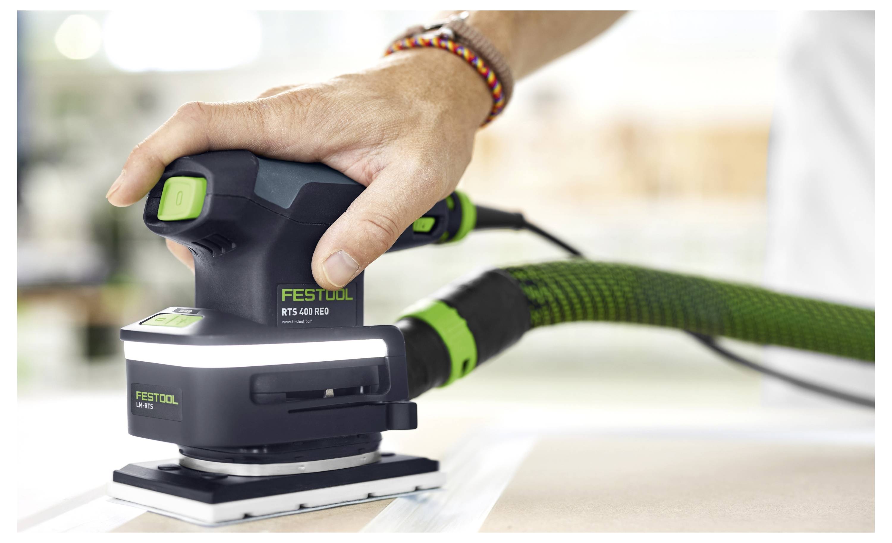 En person använder en Festool RTS 400 REQ slipmaskin med en ansluten dammsugare på en plan yta, med fokus på effektiv dammutsugning.